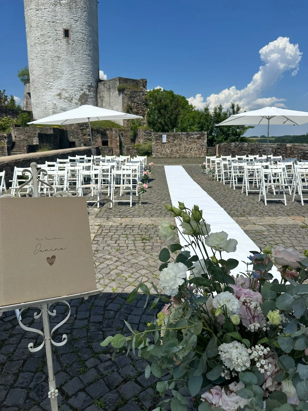 Eine Hochzeit im Freien vor einer alten Burg mit weißen Stühlen, einem roten Teppich, Blumen und Sonnenschirmen bei schönem Wetter.