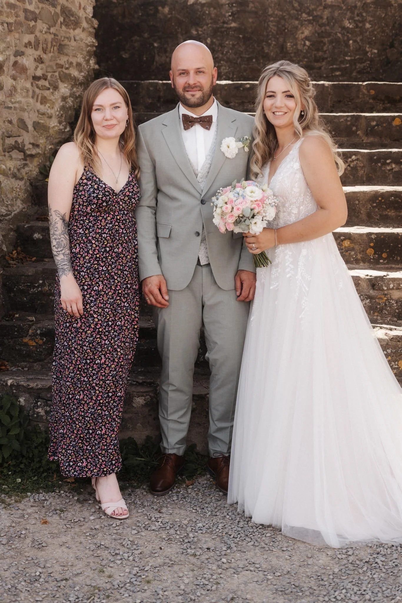 Ein Brautpaar steht auf einer Steintreppe, die Braut trägt ein weißes Hochzeitskleid und hält einen Blumenstrauß, der Bräutigam trägt einen hellgrauen Anzug, ein kariertes Fliegen, und hat einen Blumenanstecker am Revers, daneben steht eine Frau in e