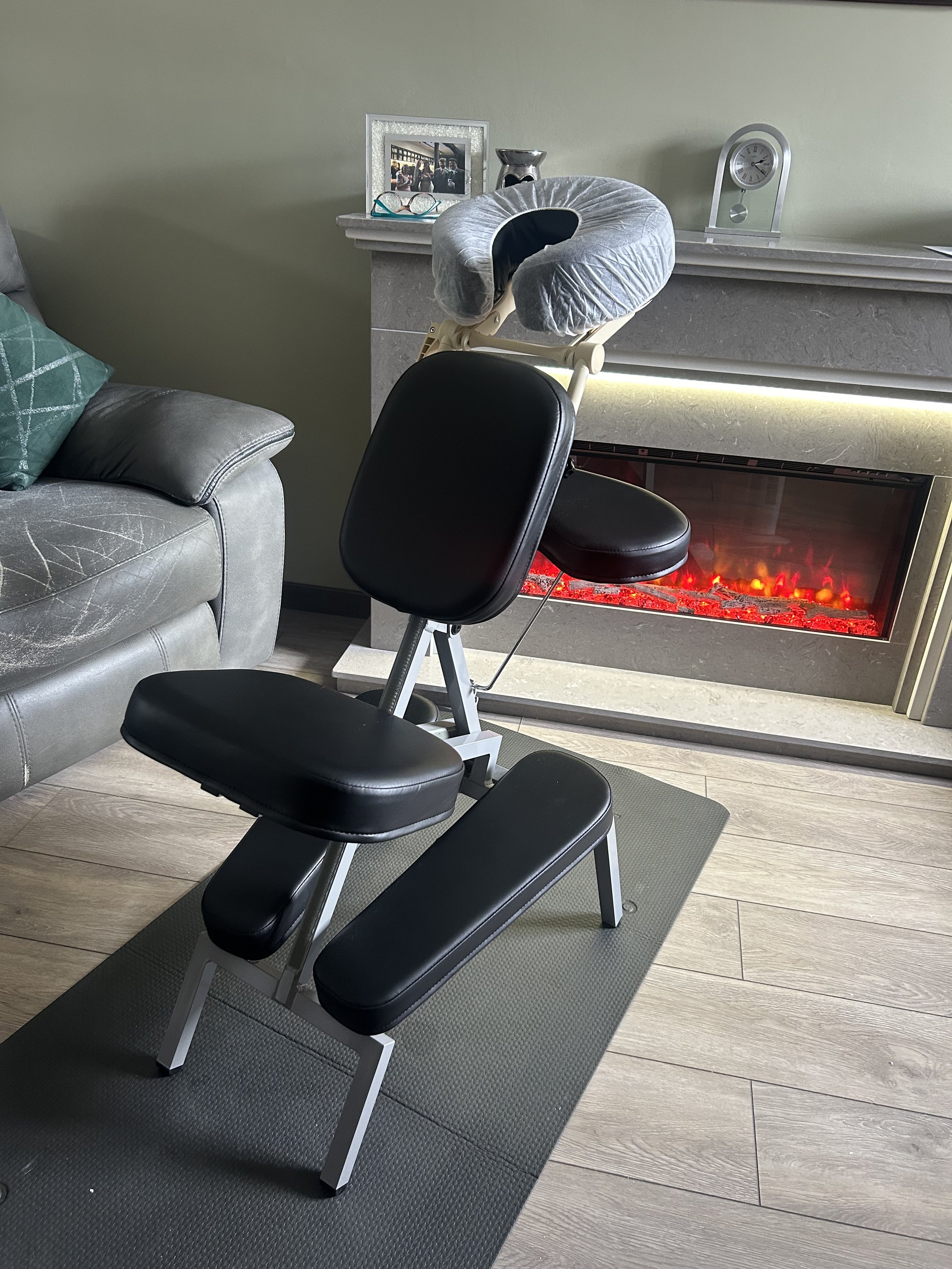 A black workout bench positioned on a gray exercise mat in front of a fireplace with a decorative clock on the mantel. To the left, part of a gray sofa with a teal pillow is visible. Behind the bench, there is a gray wall with a white mantel that displays a framed photo, a small silver sculpture, a pair of glasses, and a clock.
