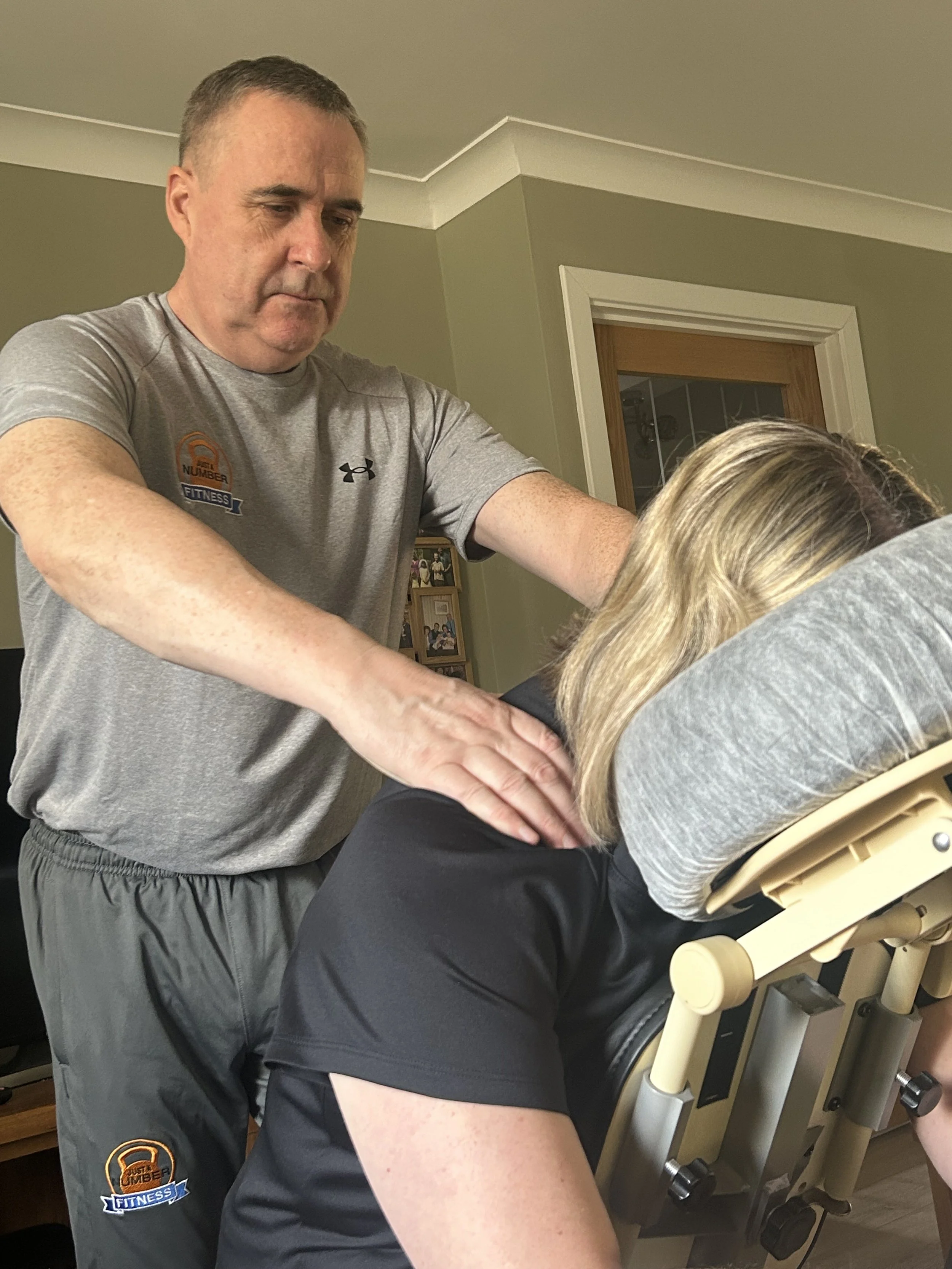 A person receiving physical therapy or chiropractic treatment from a male therapist in a clinical setting, with a treatment table and framed photographs in the background.