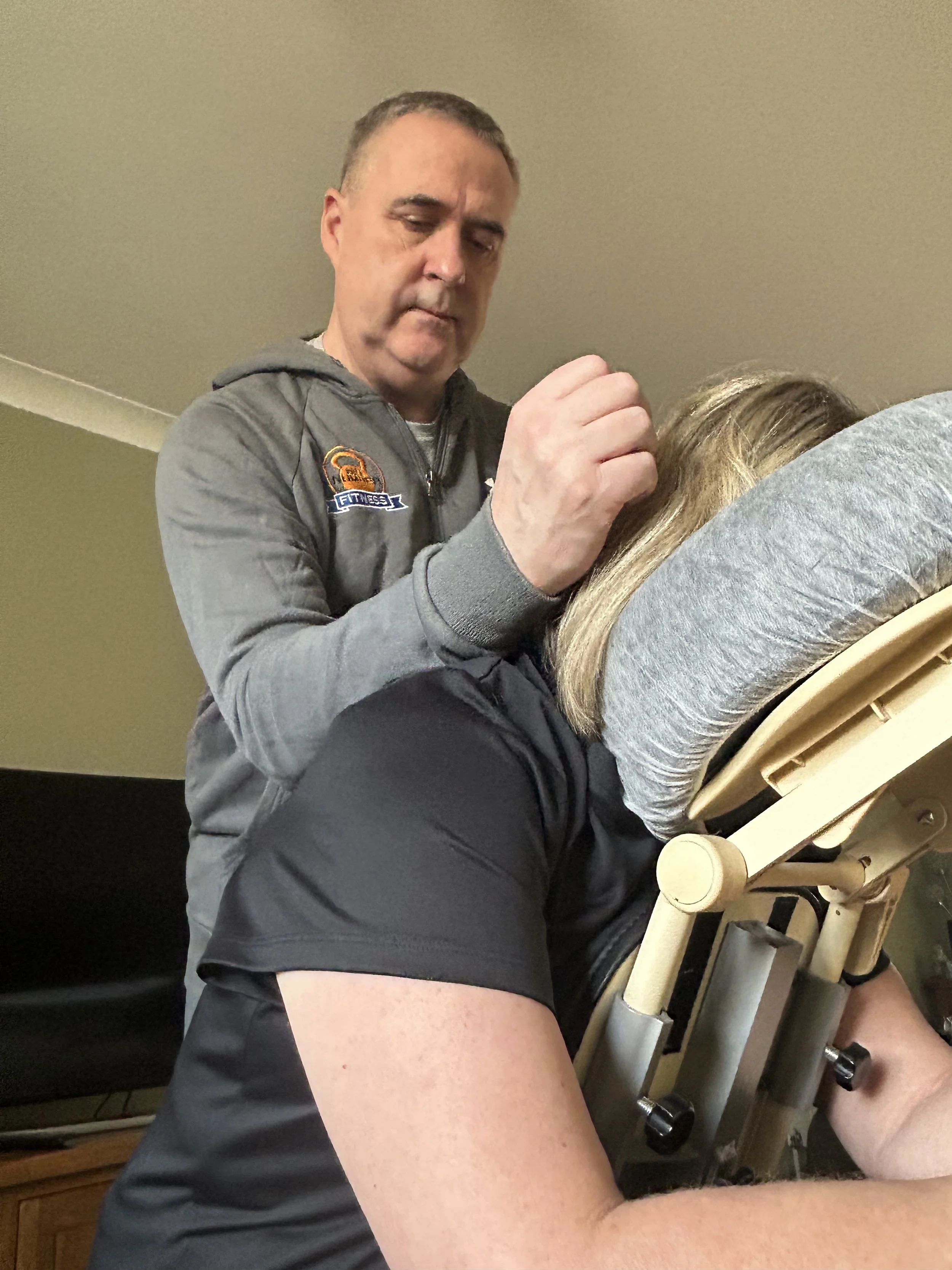 A person wearing a gray hoodie and black shorts sitting on a wooden massage table, receiving a massage, with another person not visible.