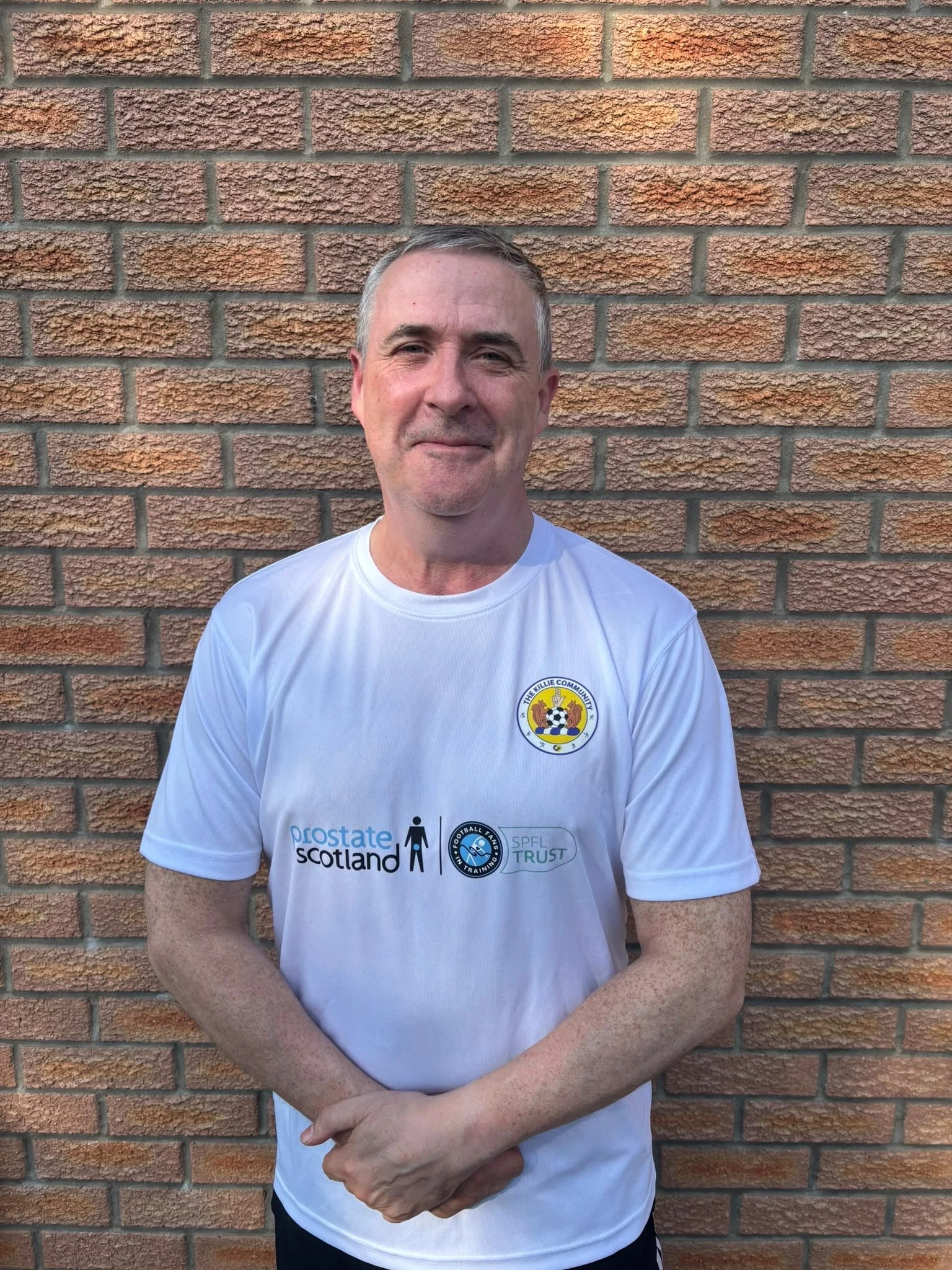 A middle-aged man with short gray hair and light skin standing outdoors against a brick wall, wearing a white sports jersey with multiple logos, including Postcode Scotland and SPFL Trust.