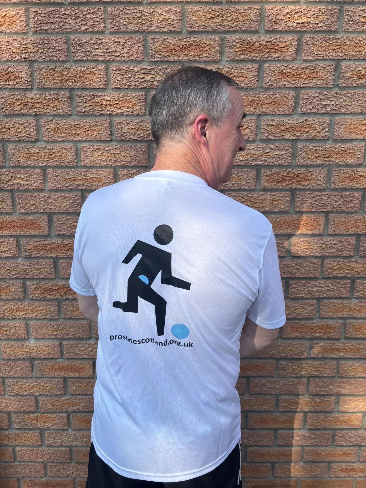 A man standing with his back to the camera, wearing a white T-shirt with a logo and website for prostate cancer awareness, standing against a brick wall.