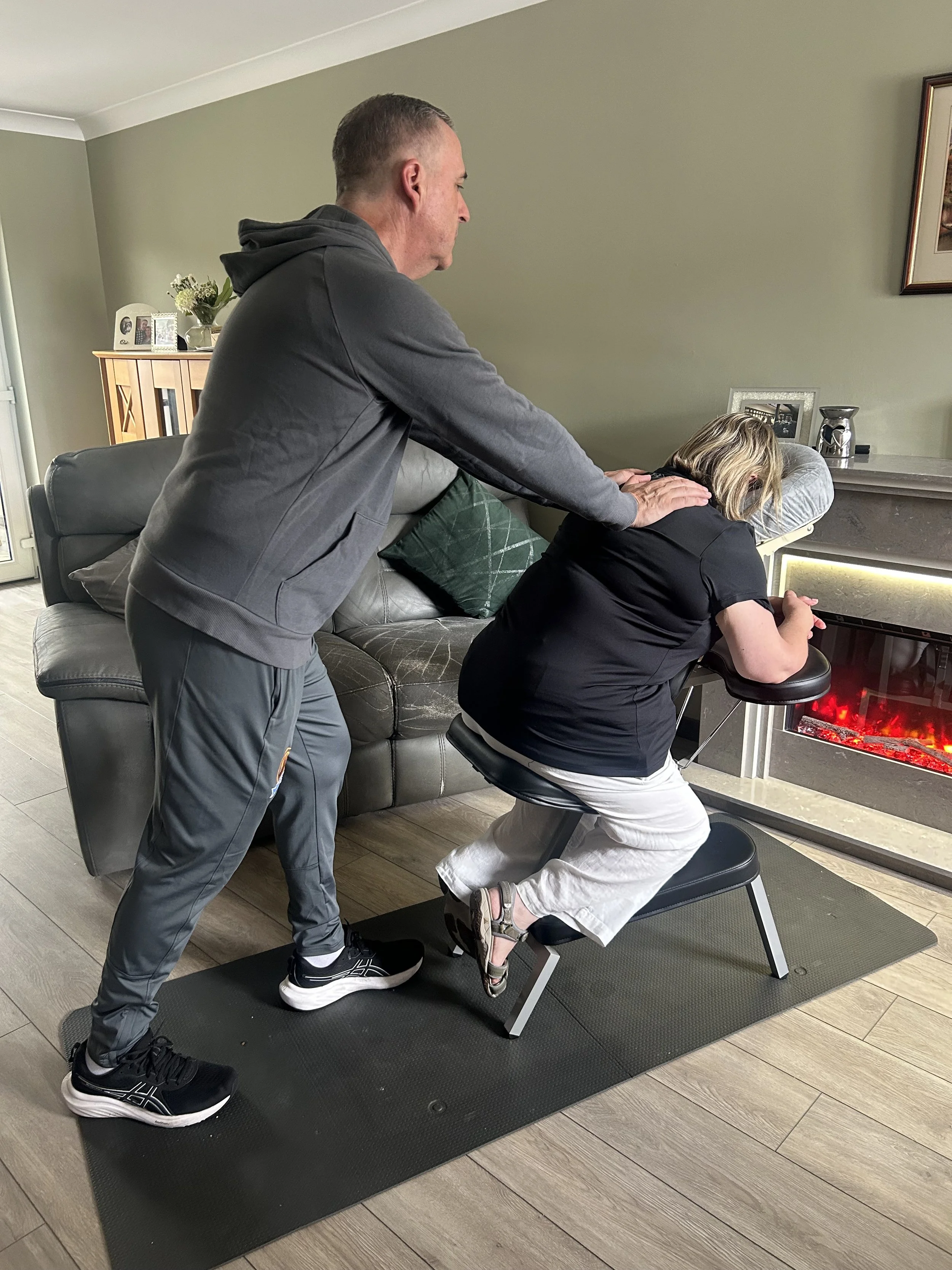 A man providing a back massage to a woman seated in a massage chair in a living room with a fireplace, decorated with family photos and flowers.