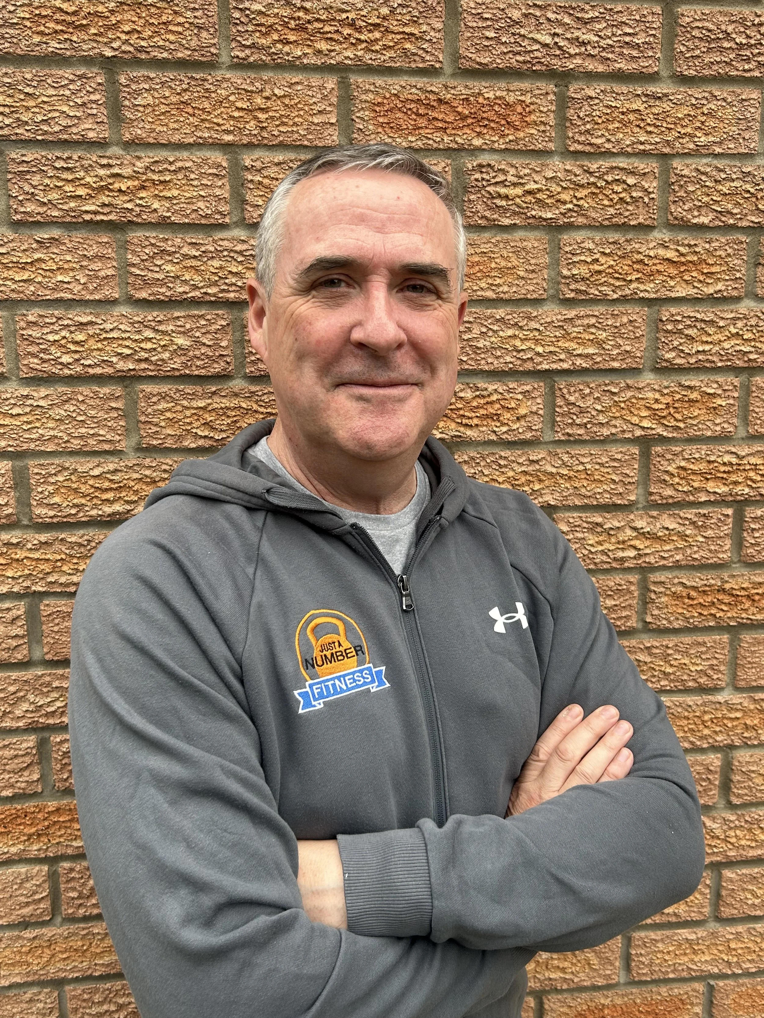A middle-aged man with short gray hair, wearing a dark gray jacket with a fitness logo and an Under Armour logo, standing against a brick wall. He has his arms crossed and is smiling.