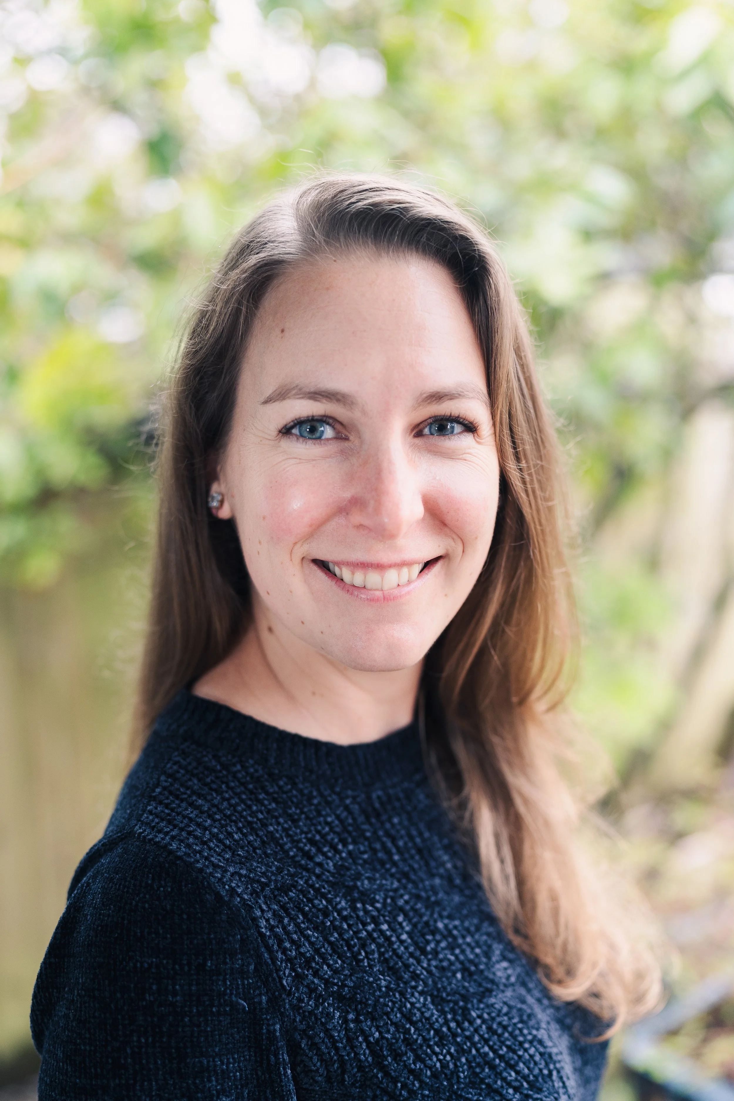 Close-up portrait of a woman with long brown hair, blue eyes, and earrings, smiling outdoors with a blurred green background.