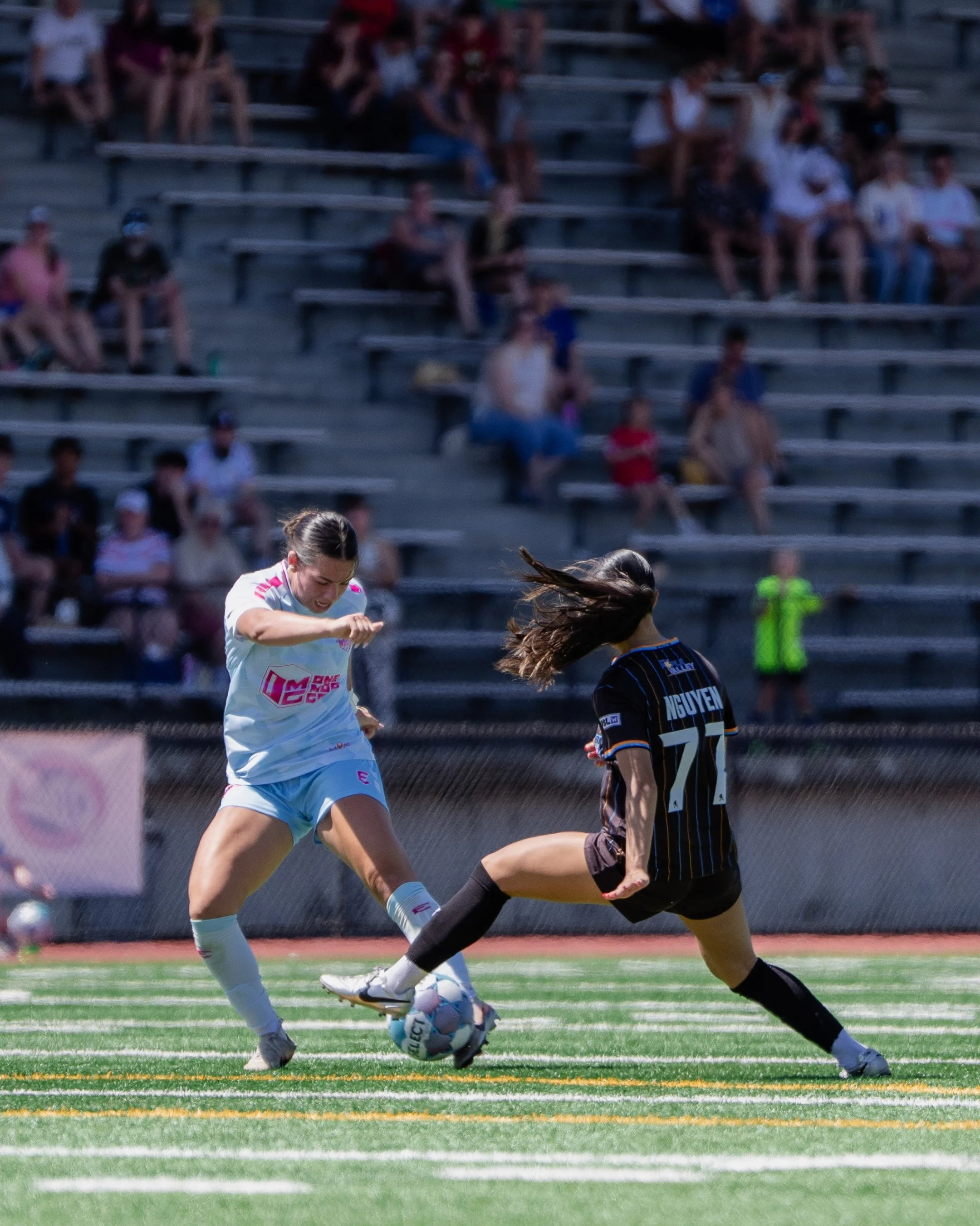 Two female soccer players competing for the ball during a match on a green field, with spectators sitting in the bleachers in the background.