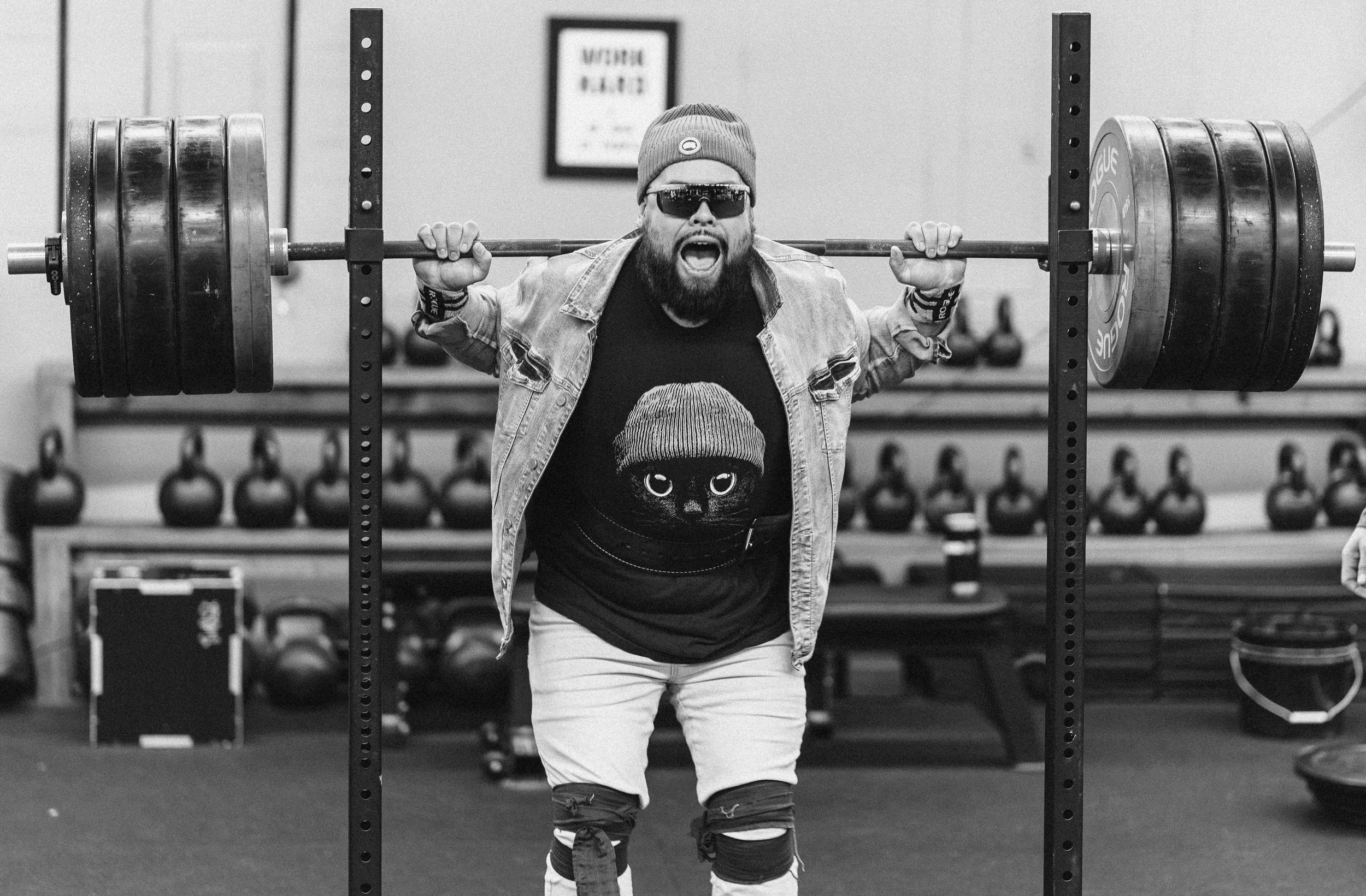 A bearded man wearing sunglasses, a beanie, a denim jacket, and a t-shirt with a cartoon face, is lifting a heavily loaded barbell in a gym. He appears to be shouting or cheering.