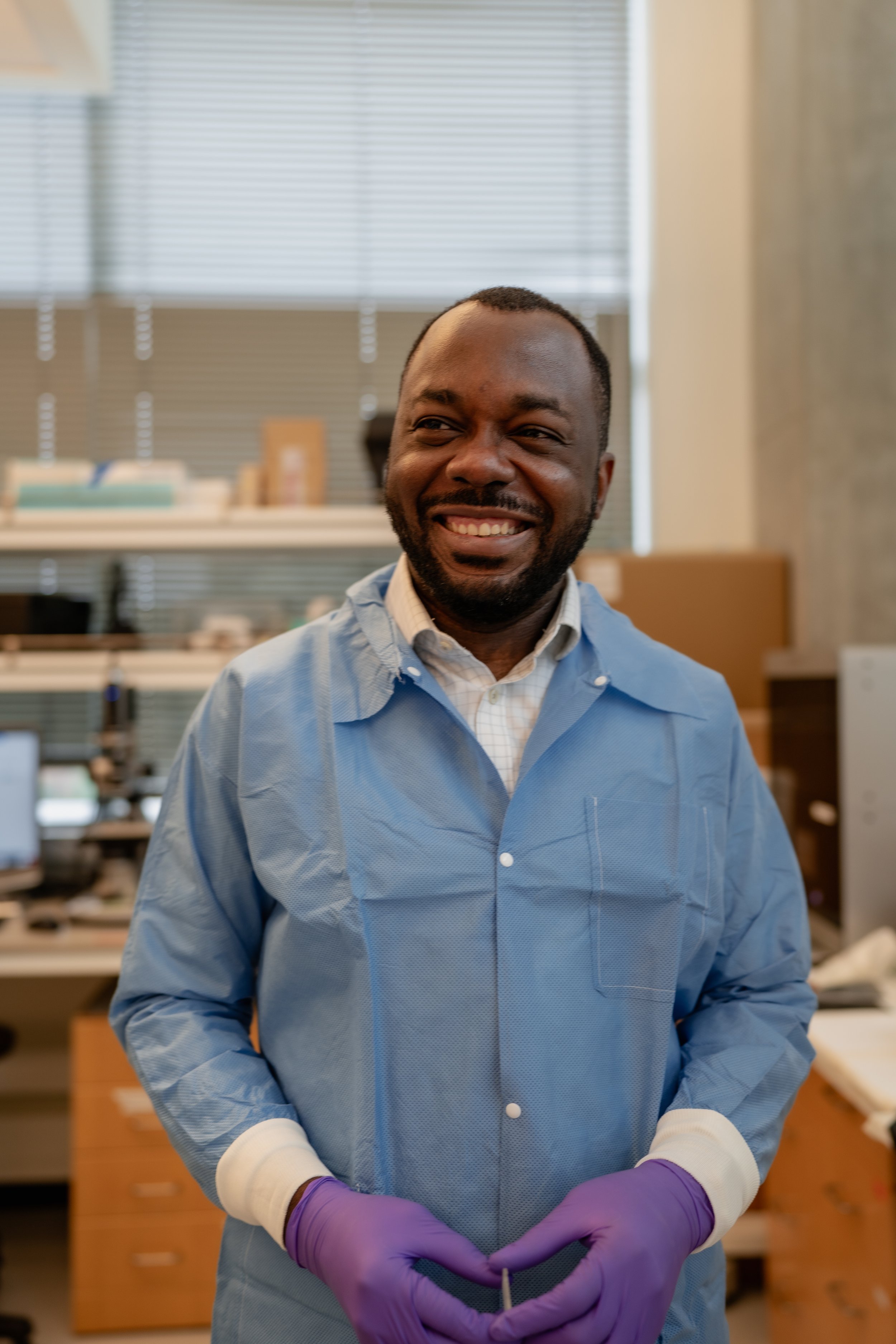 A smiling man in a blue lab coat and purple gloves standing in a laboratory environment.