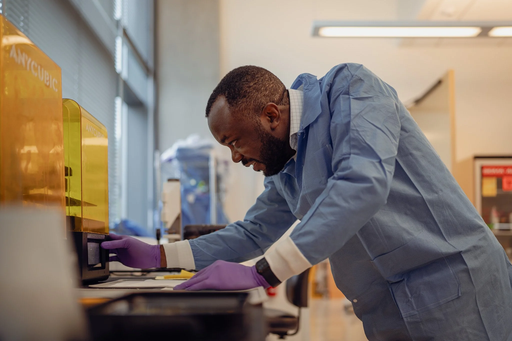 A scientist in a laboratory wearing purple gloves and a blue lab coat, working with scientific equipment on a lab bench, with a focus on his side profile.