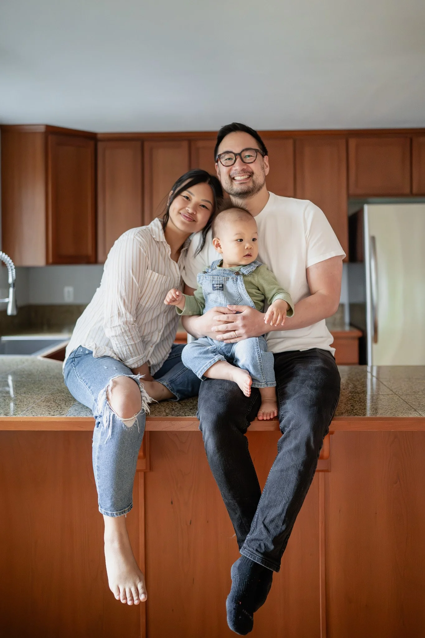 A happy diverse family sitting on a kitchen countertop, smiling at the camera. The family includes a woman, a man, and a young child.