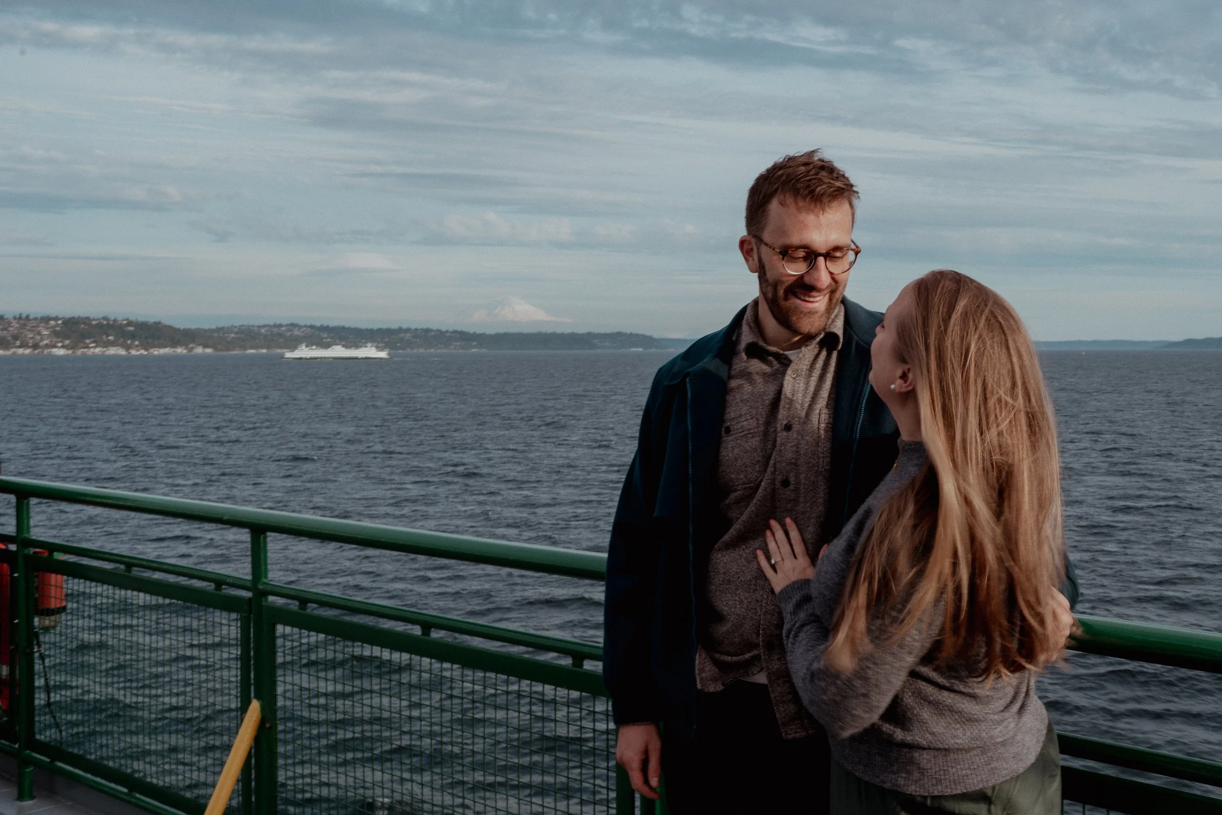 A man and woman standing on a boat deck near a green railing, smiling and looking at each other with water and a distant shore in the background.