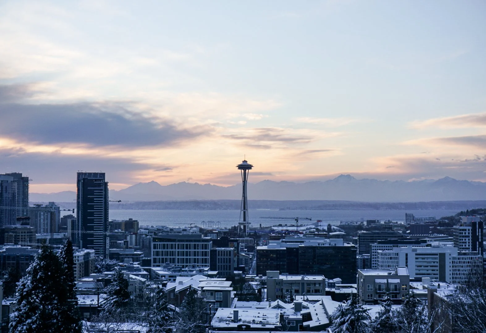 A cityscape of Seattle during winter with snow-covered buildings, the Space Needle in the center, Mount Rainier in the background, and a partly cloudy sky.