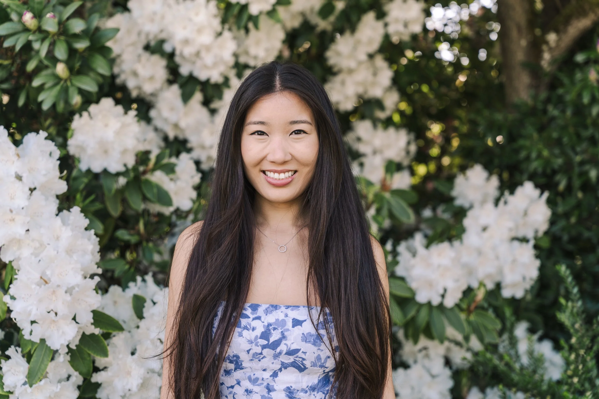 A smiling woman with long dark hair standing outdoors in front of white flowering bushes.