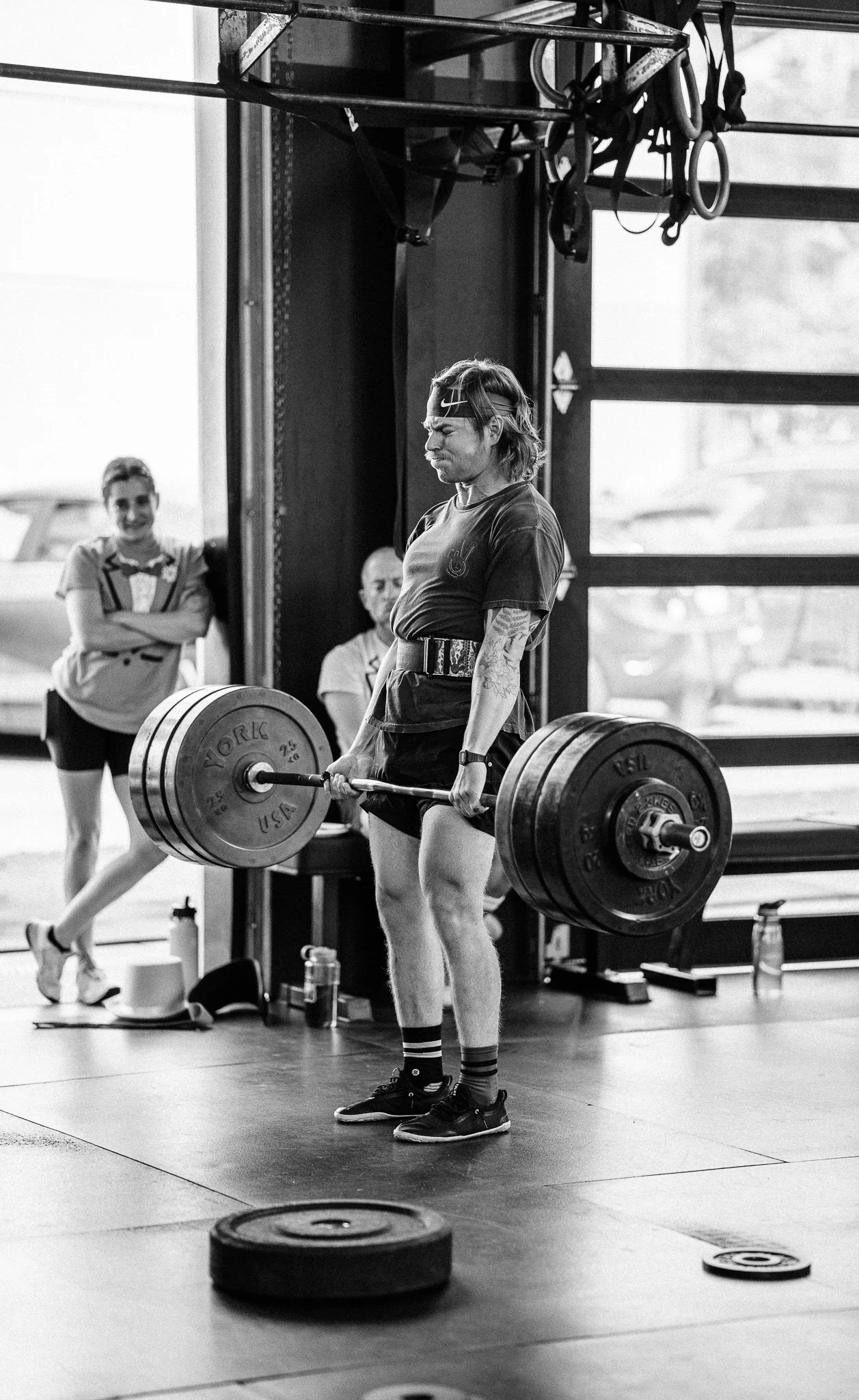 A man lifting a barbell with weight plates during a workout at the gym while two women observe in the background.