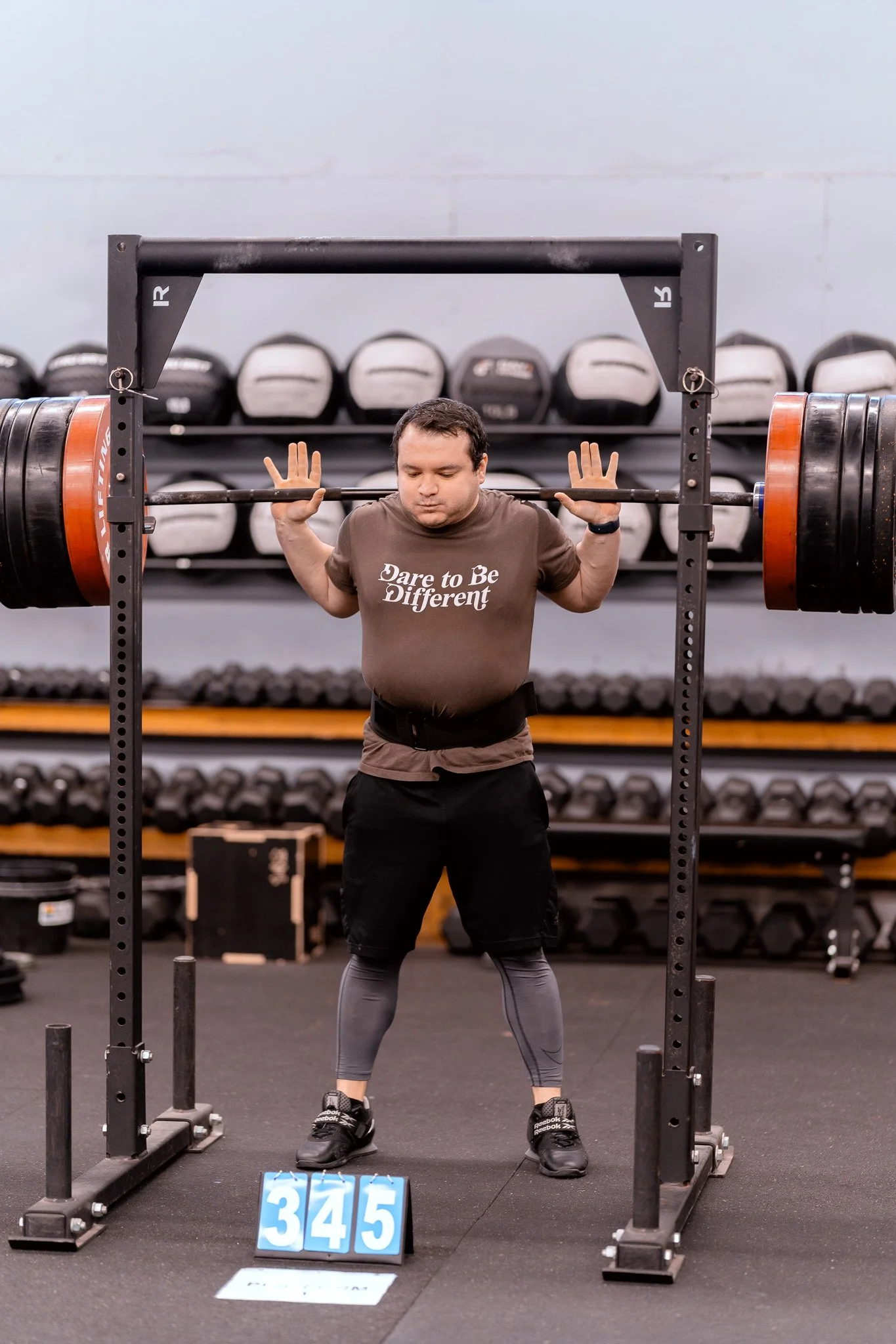 Man wearing a brown T-shirt with the words "Dare to Be Different" lifts a heavy barbell during weightlifting at a gym.