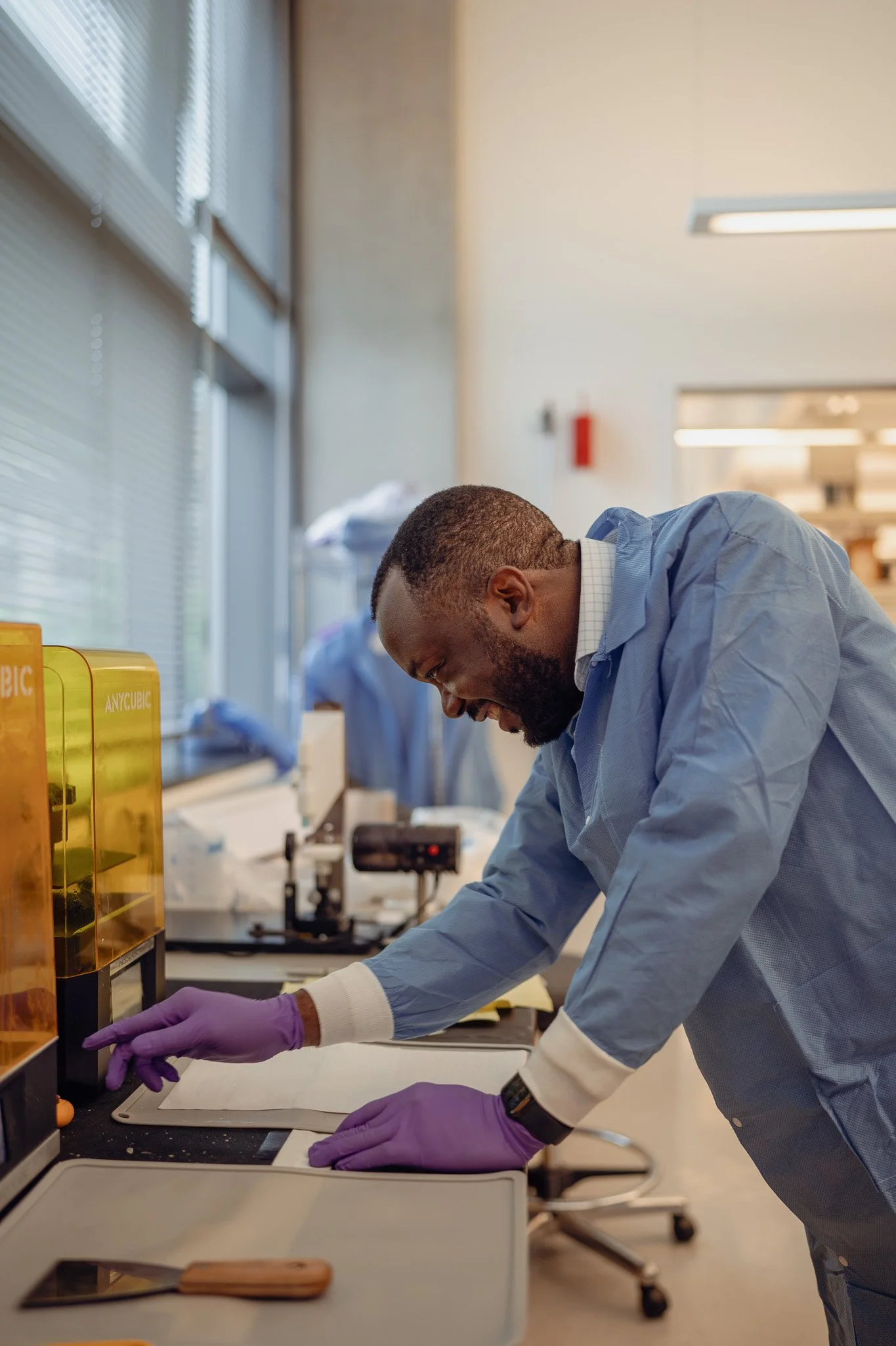 A scientist in a lab coat and purple gloves leaning over a work surface, smiling and working with scientific equipment in a laboratory with large windows.