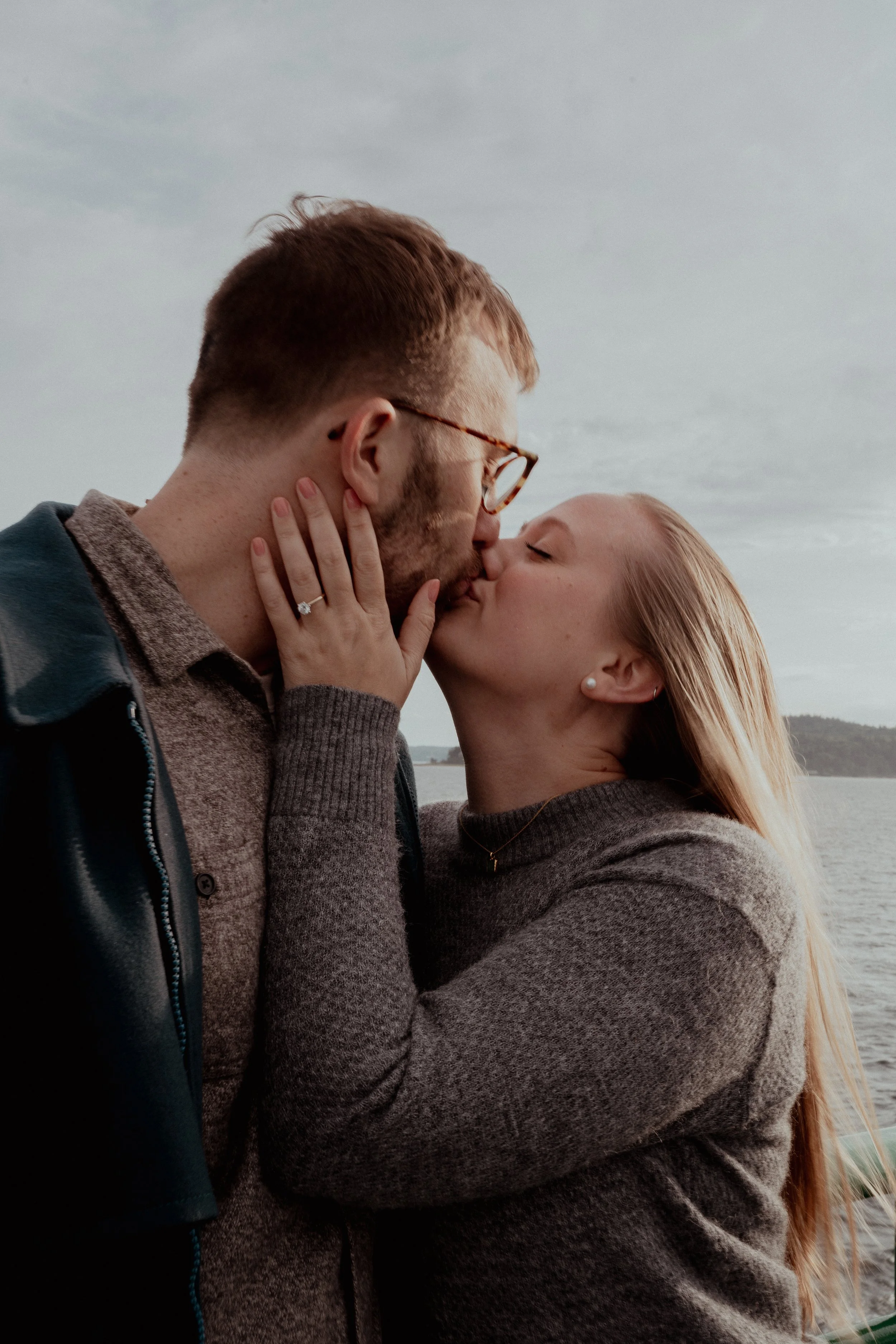 A couple is kissing near a body of water, with a scenic view of the lake and distant hills in the background.