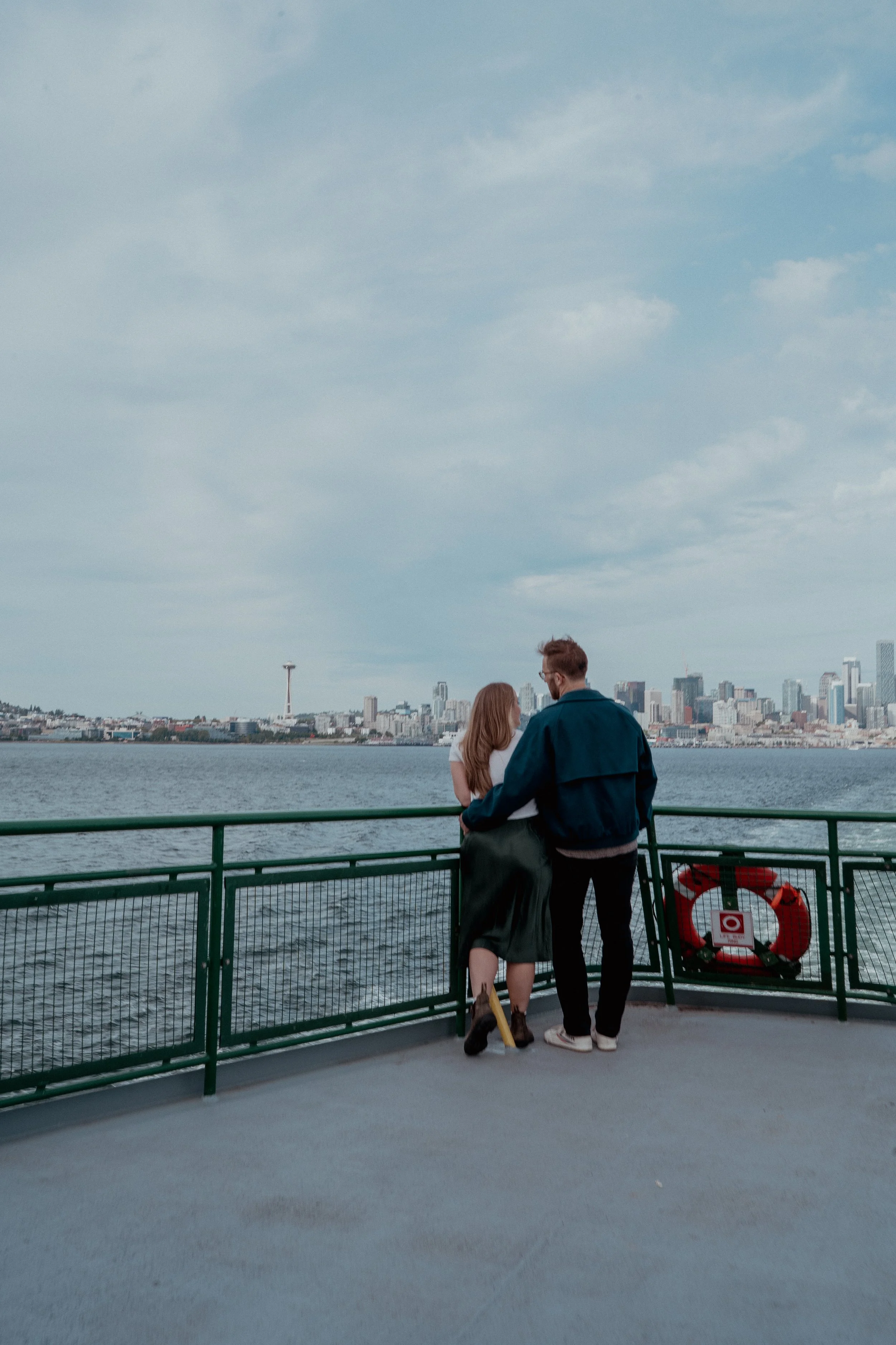A couple standing on a boat deck, overlooking a city skyline across the water, with a ferris wheel and skyscrapers in the background.
