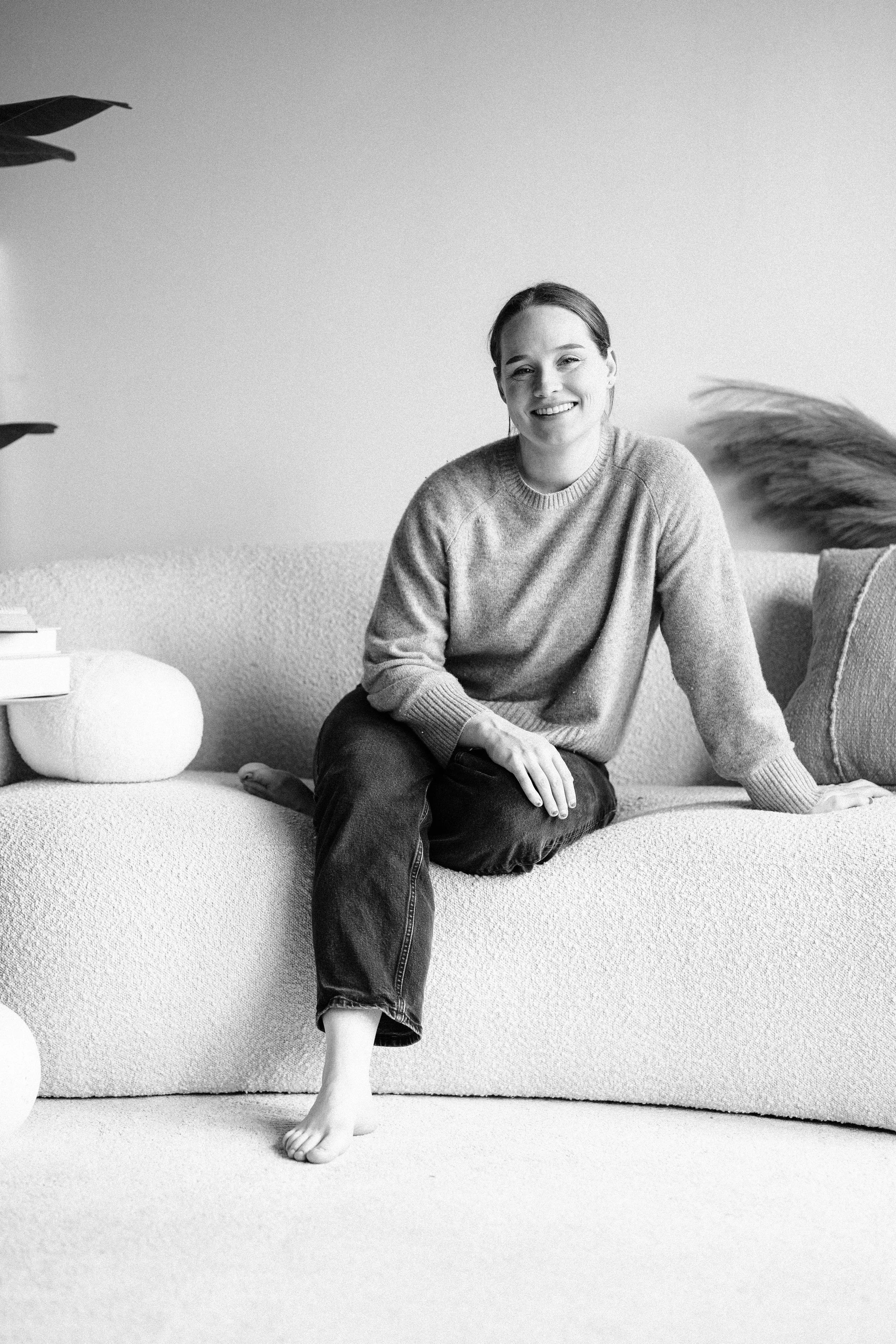 A woman smiling while sitting on a textured sofa in a living room, wearing a sweater and jeans.