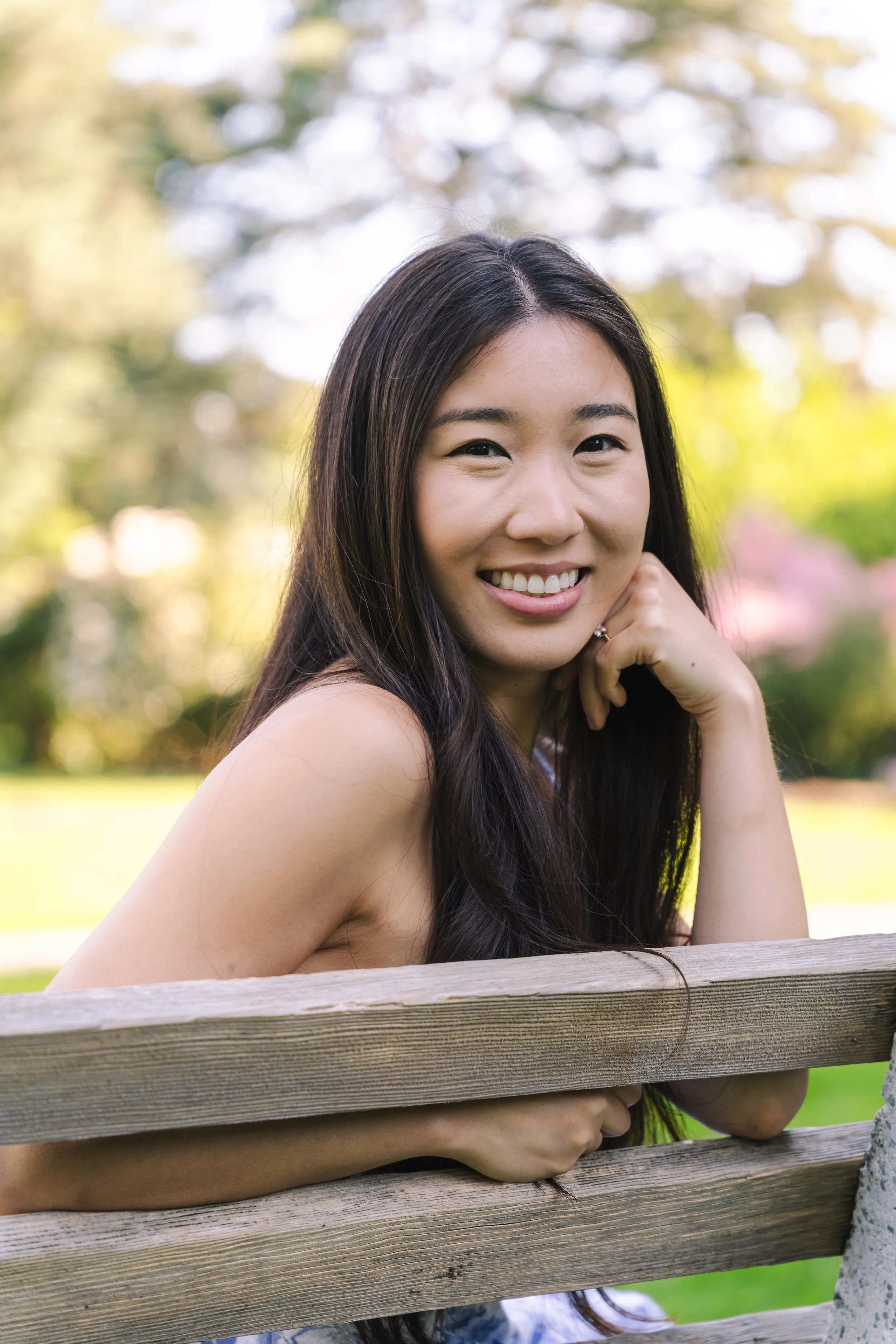 A young woman with long dark hair smiling and resting her chin on her hand, standing outdoors behind a wooden fence with a blurred background of trees and sunlight.