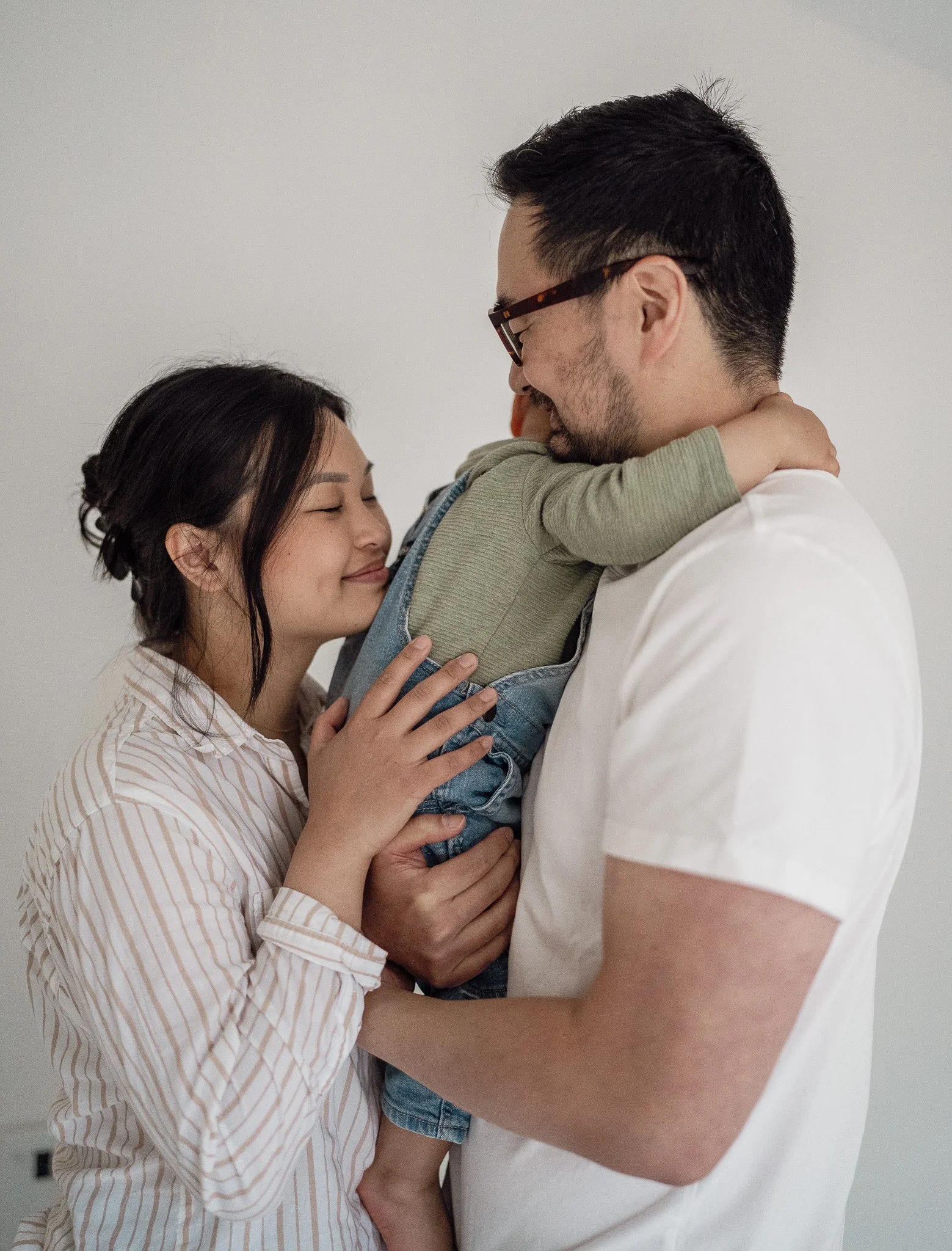A young family hugging, with the mother and father holding their small child between them, all smiling contentedly.