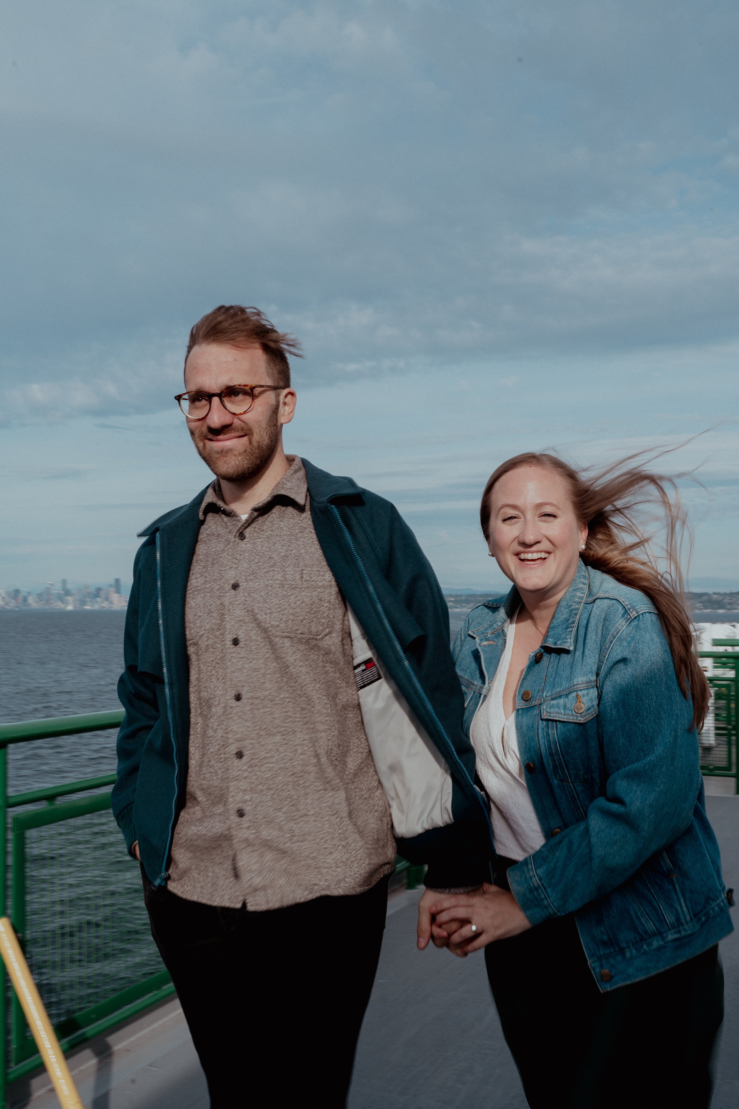 A man and a woman standing outdoors on a boat deck, holding hands with a city skyline and water in the background, both smiling and enjoying a windy day.