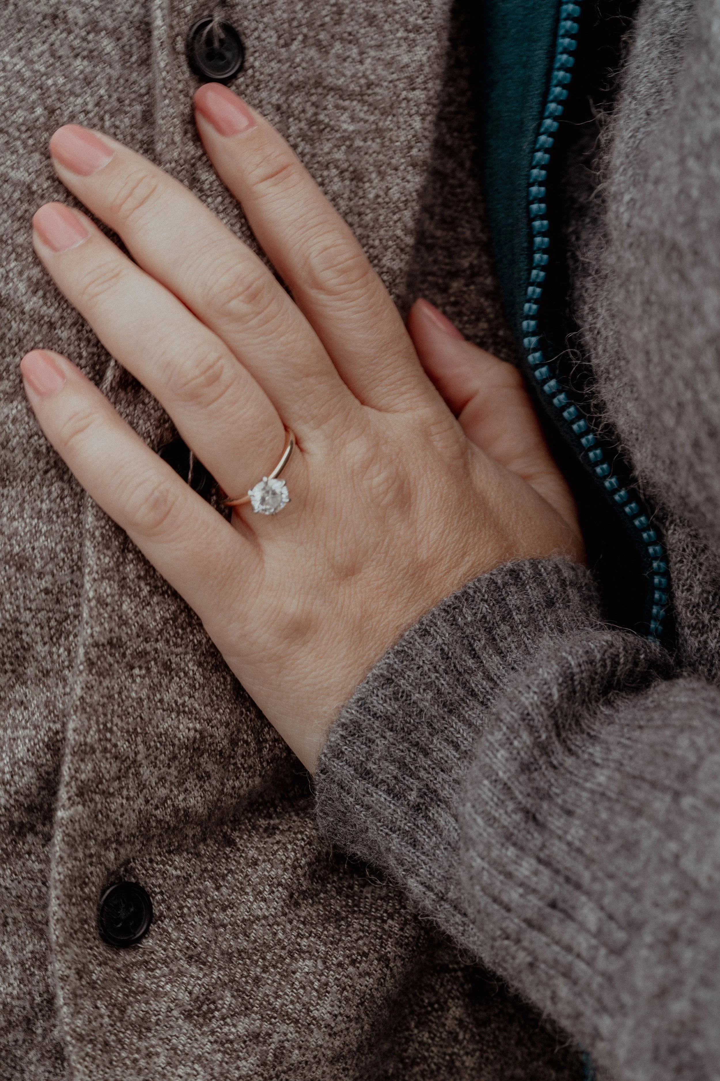 Close-up of a woman wearing a ring with a large gemstone on her left ring finger, resting her hand on her chest, with part of her beige and blue clothing visible.