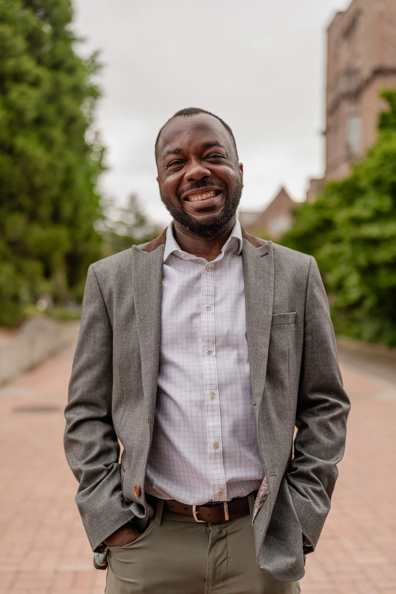 Smiling man in a grey blazer and light shirt standing outdoors on a brick pathway with greenery and historic buildings in the background.