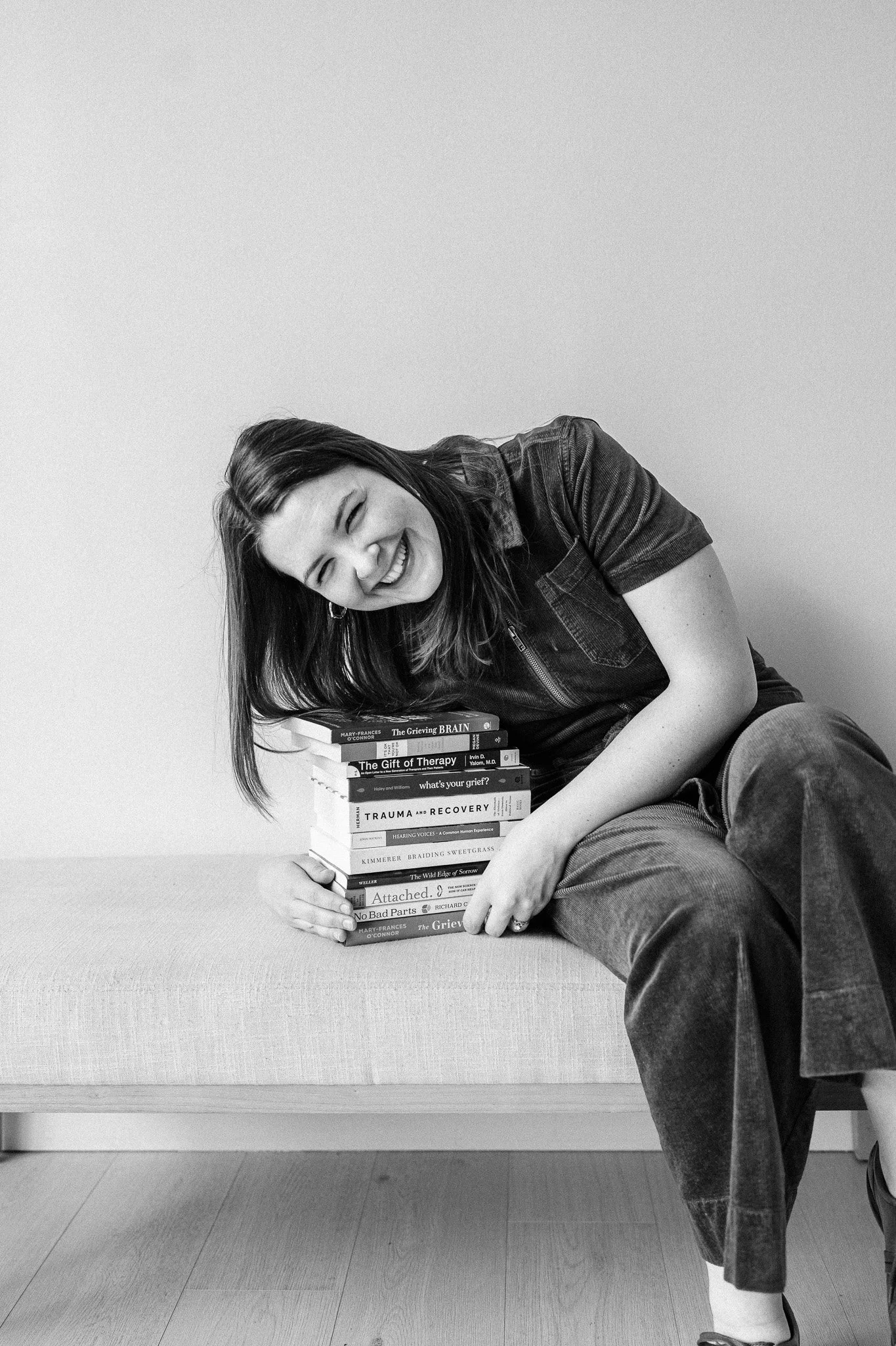 A woman with long dark hair, wearing a dark denim jacket and jeans, is sitting on the floor, smiling with her eyes closed. She is leaning on a stack of books, which include titles like 'The Grieving Brain,' 'The Gift of Therapy,' and 'Trauma and Recovery.' The background is a plain light-colored wall.