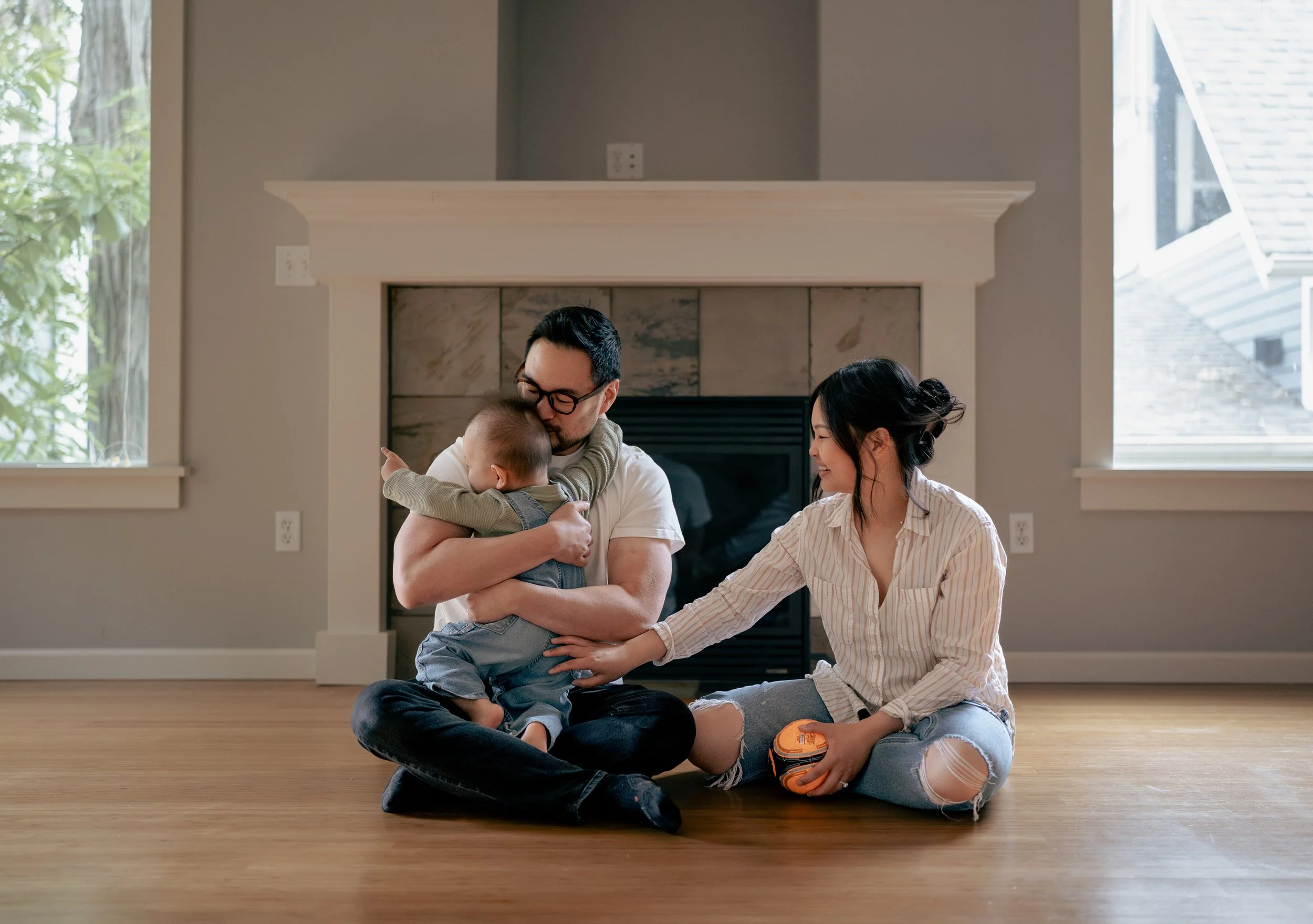 A family sitting on a wooden floor in front of a fireplace, with a man holding a young child and a woman smiling and reaching towards the child.