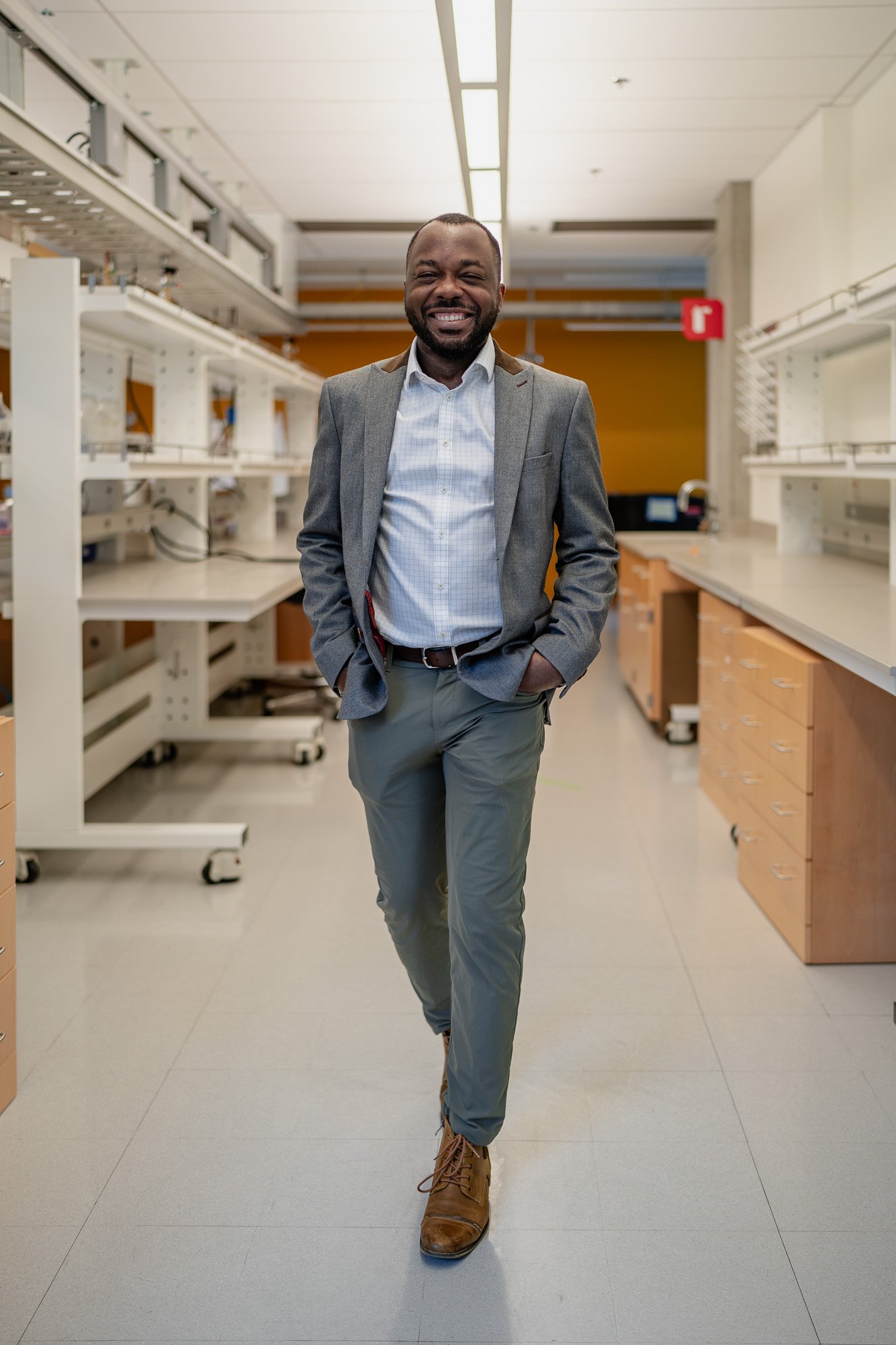 A smiling man in a gray blazer, checkered shirt, gray pants, and brown boots walking through a laboratory or workspace with shelves and drawers.