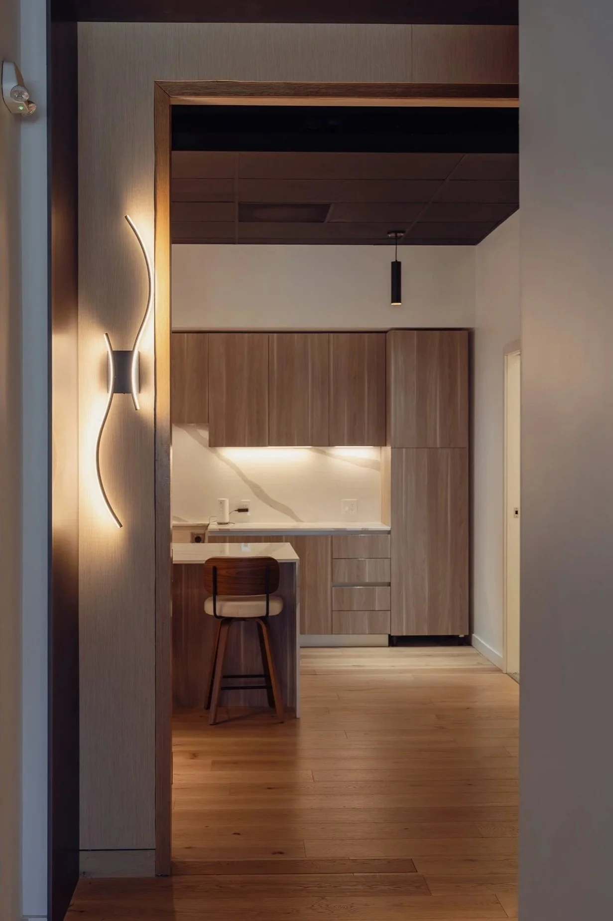 Interior view of a modern kitchen with wooden cabinets, a small breakfast bar with a chair, and wall-mounted lamps. The flooring is wooden, and the ceiling has a dark brown panel with a hanging light fixture.
