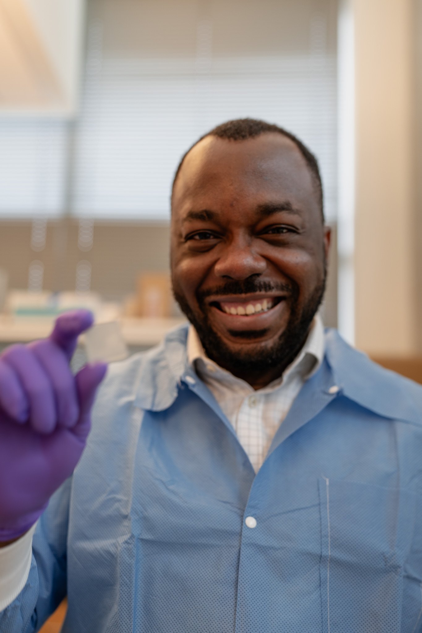 A man smiling in a laboratory, wearing a blue lab coat and purple gloves, holding a small sample container.