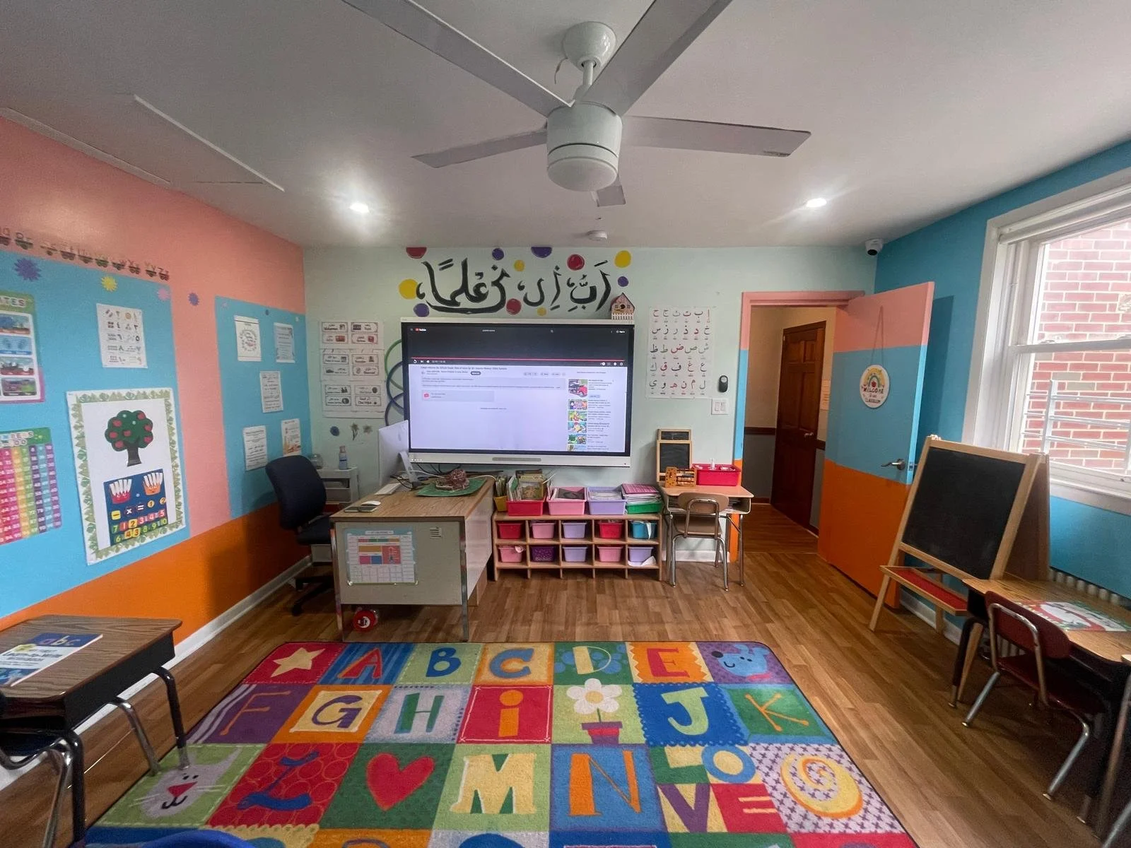 Colorful classroom with a large alphabet rug, blackboard, wall charts, and a digital whiteboard in the center. The room has pink, blue, and orange walls, a ceiling fan, and a window letting in natural light.