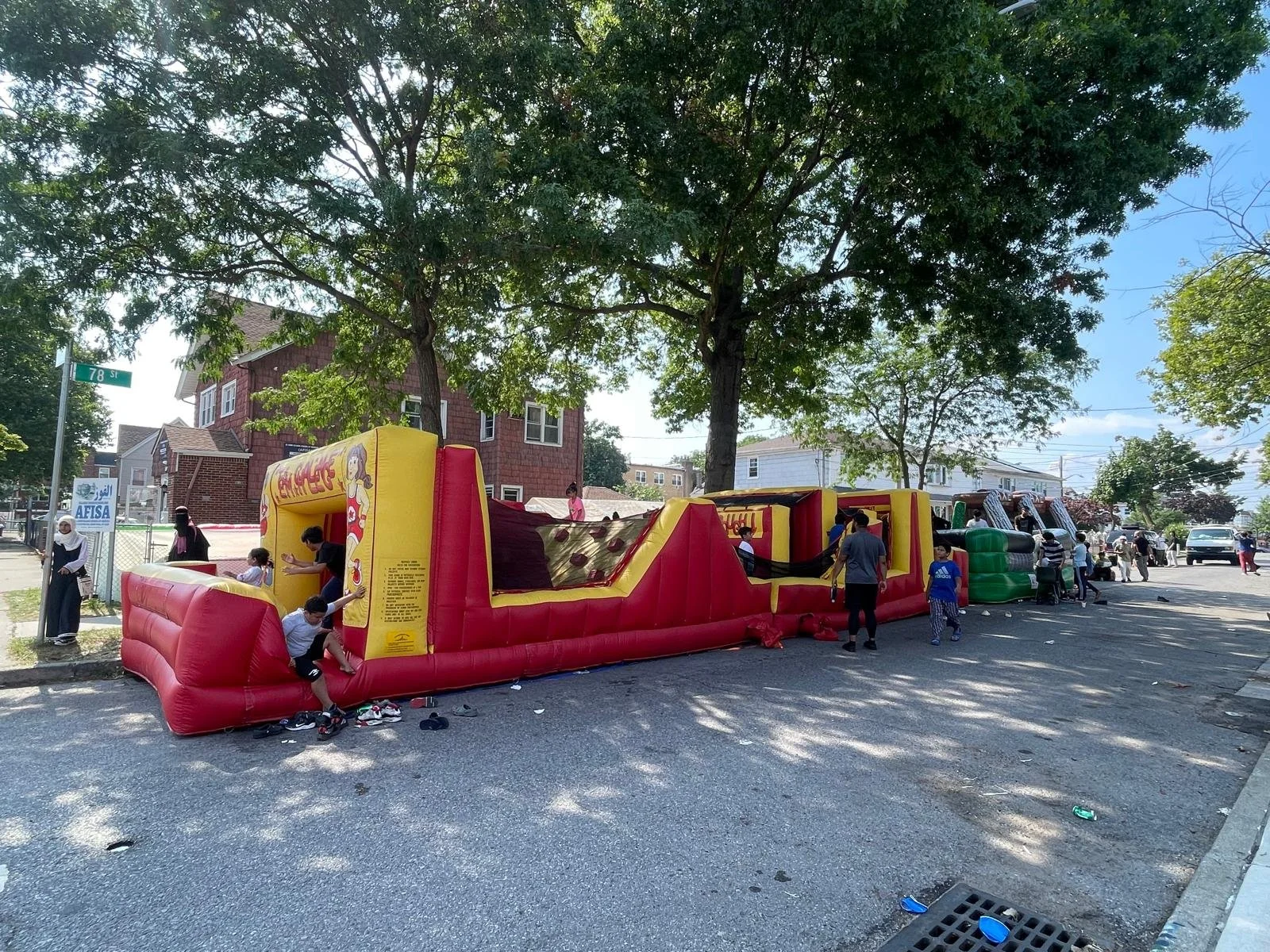 Children playing on an inflatable bounce house and slide in a neighborhood street, with trees and houses in the background.