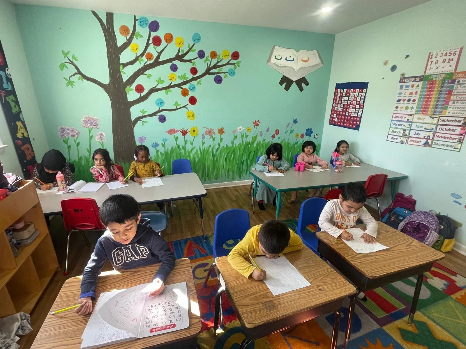 Young children seated at desks in a colorful classroom, working on writing activities. The walls are decorated with a large painted tree with colored circles on the branches, and educational posters.