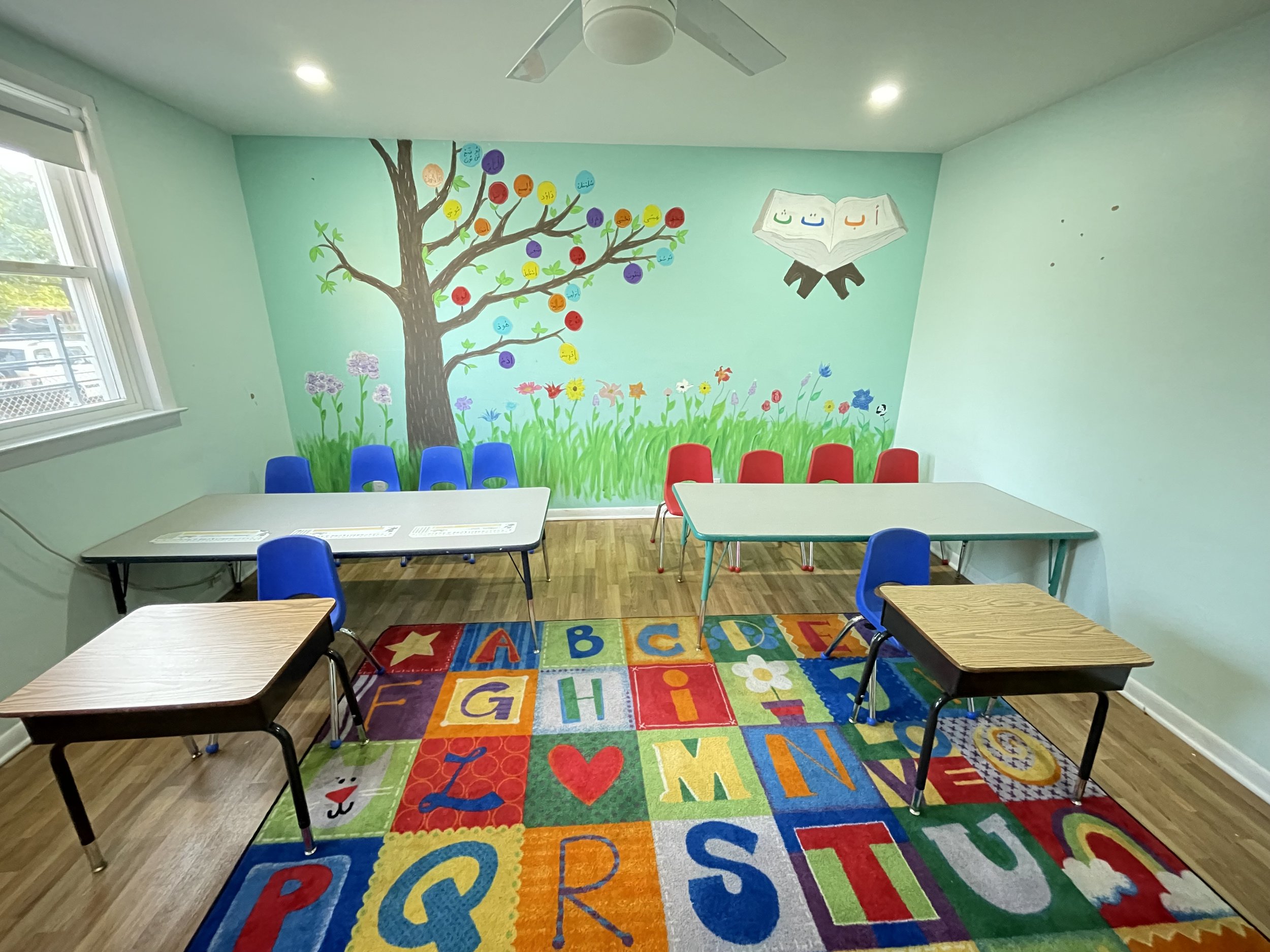 A brightly decorated classroom with a colorful alphabet rug, two long tables with chairs, and a mural of a tree with colorful circles on its branches, flowers, and an open book with Arabic letters on the wall.