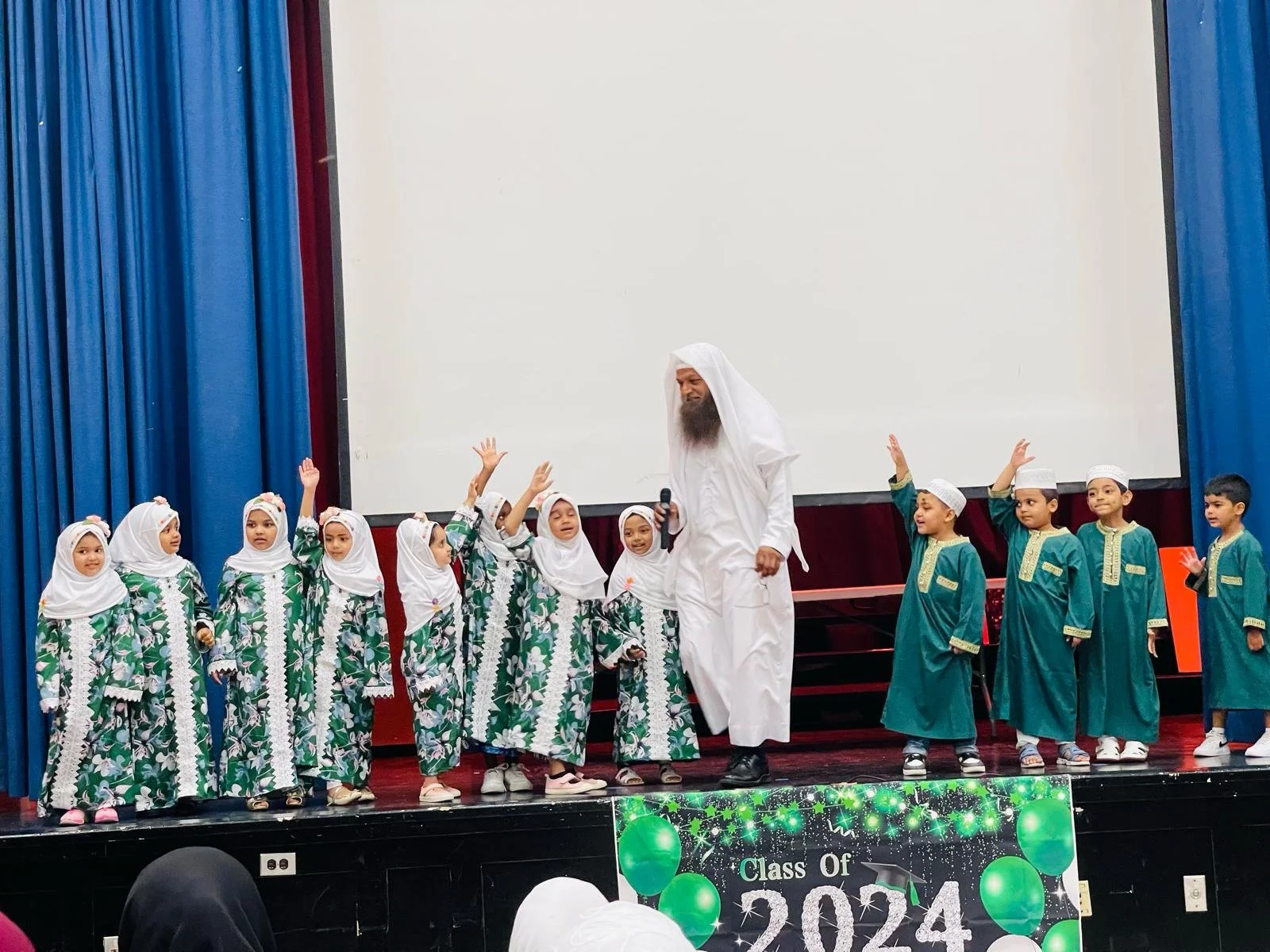 Children in traditional clothing on stage during a school graduation ceremony, with a man in white attire in the center, some children raising their hands, and a banner reading 'Class of 2024' at the front.