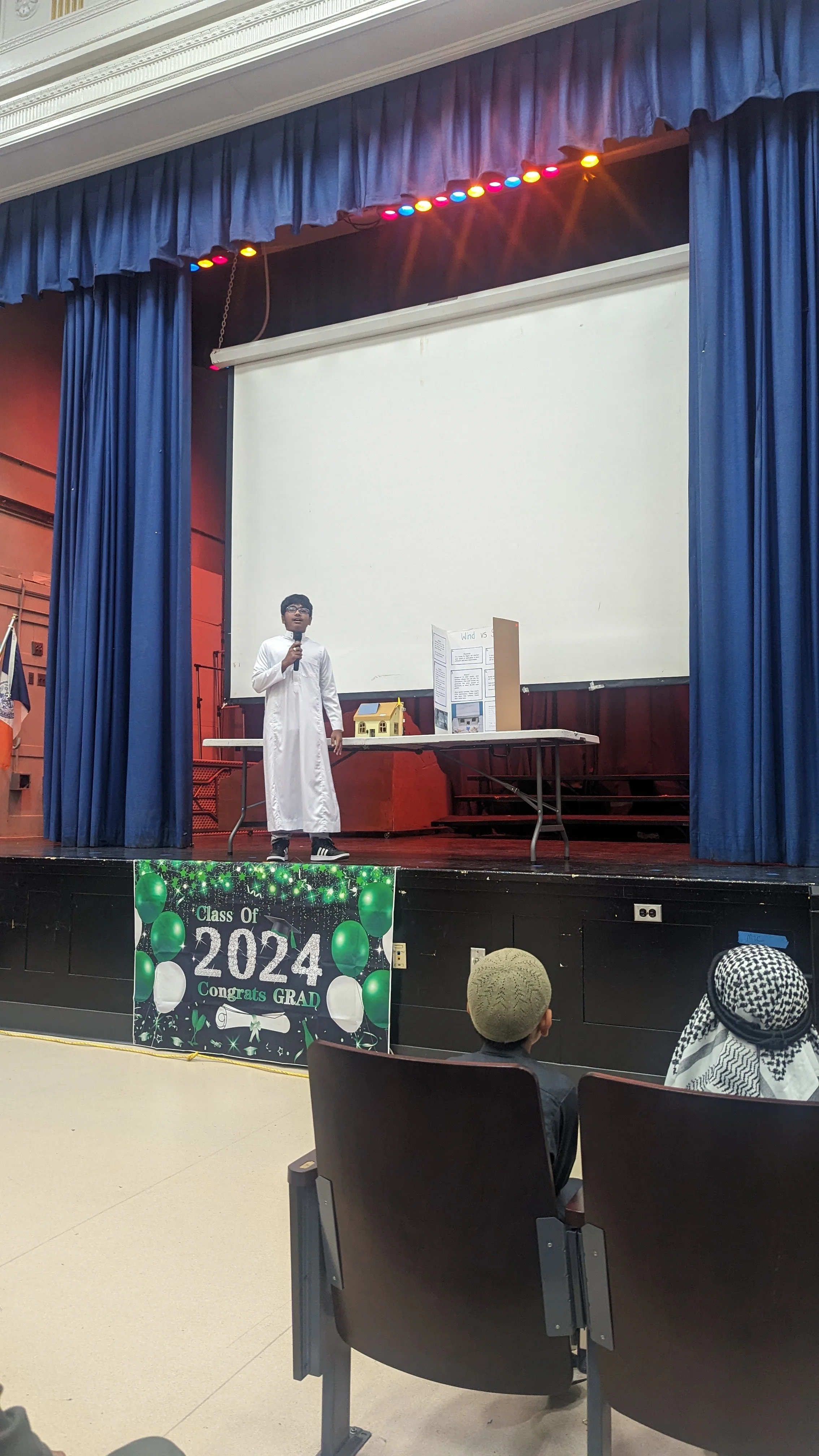 A young student in a white gown giving a presentation on a stage decorated for a graduation ceremony, with a banner reading 'Class of 2024 Congrats GRAD' and an audience of children seated in front.
