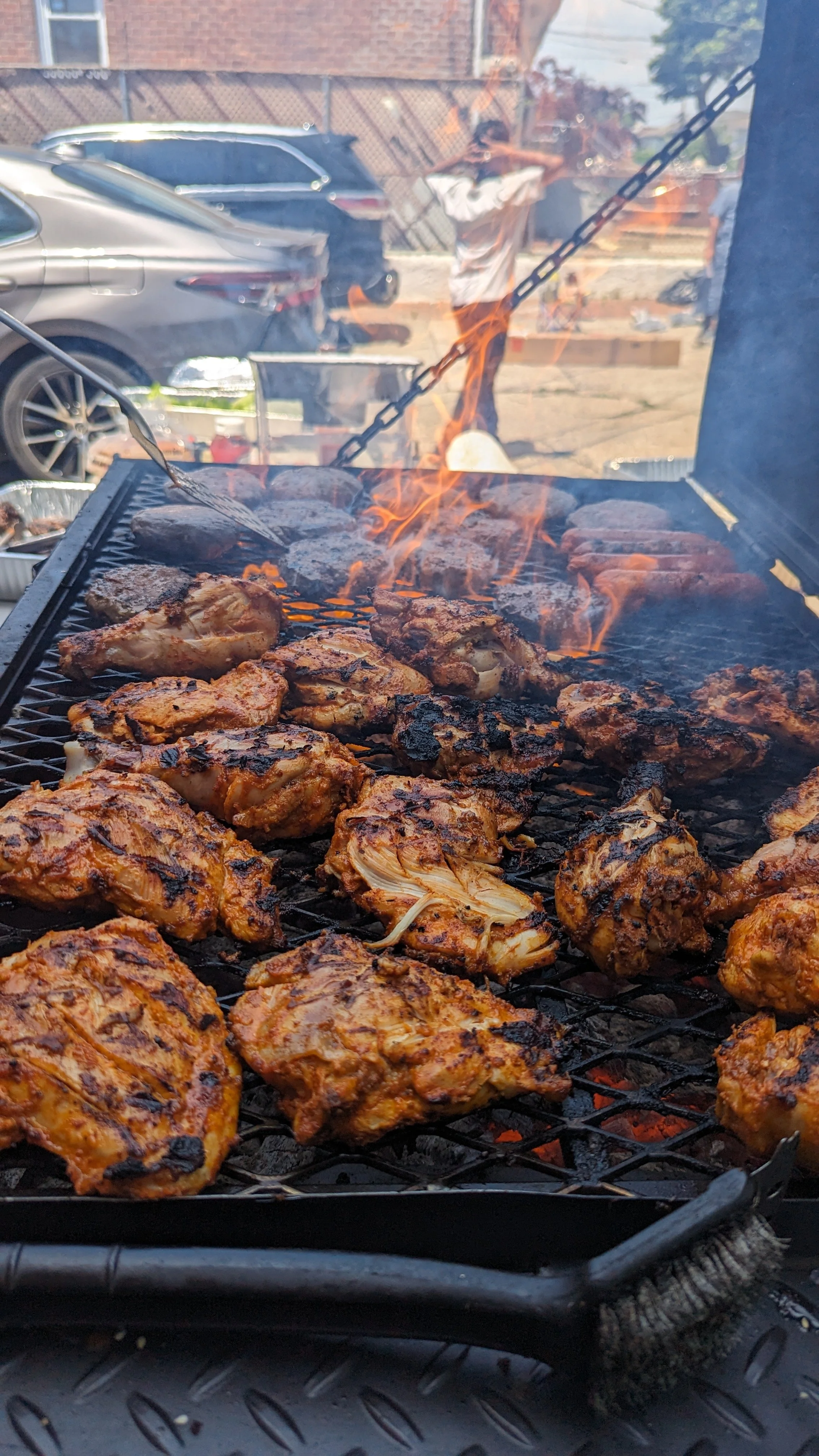 Barbecue grill with grilled chicken and burger patties, flames visible in the background, outdoor setting with cars and people.