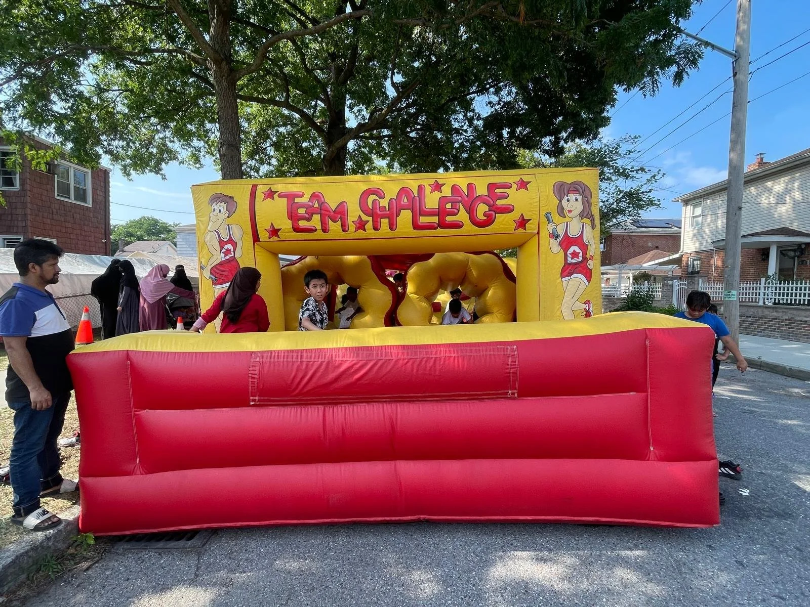 Children playing on a yellow and red inflatable bounce house with the name 'Team Challenge' and cartoon images of girls on the top panel, set outdoors on a sunny day.