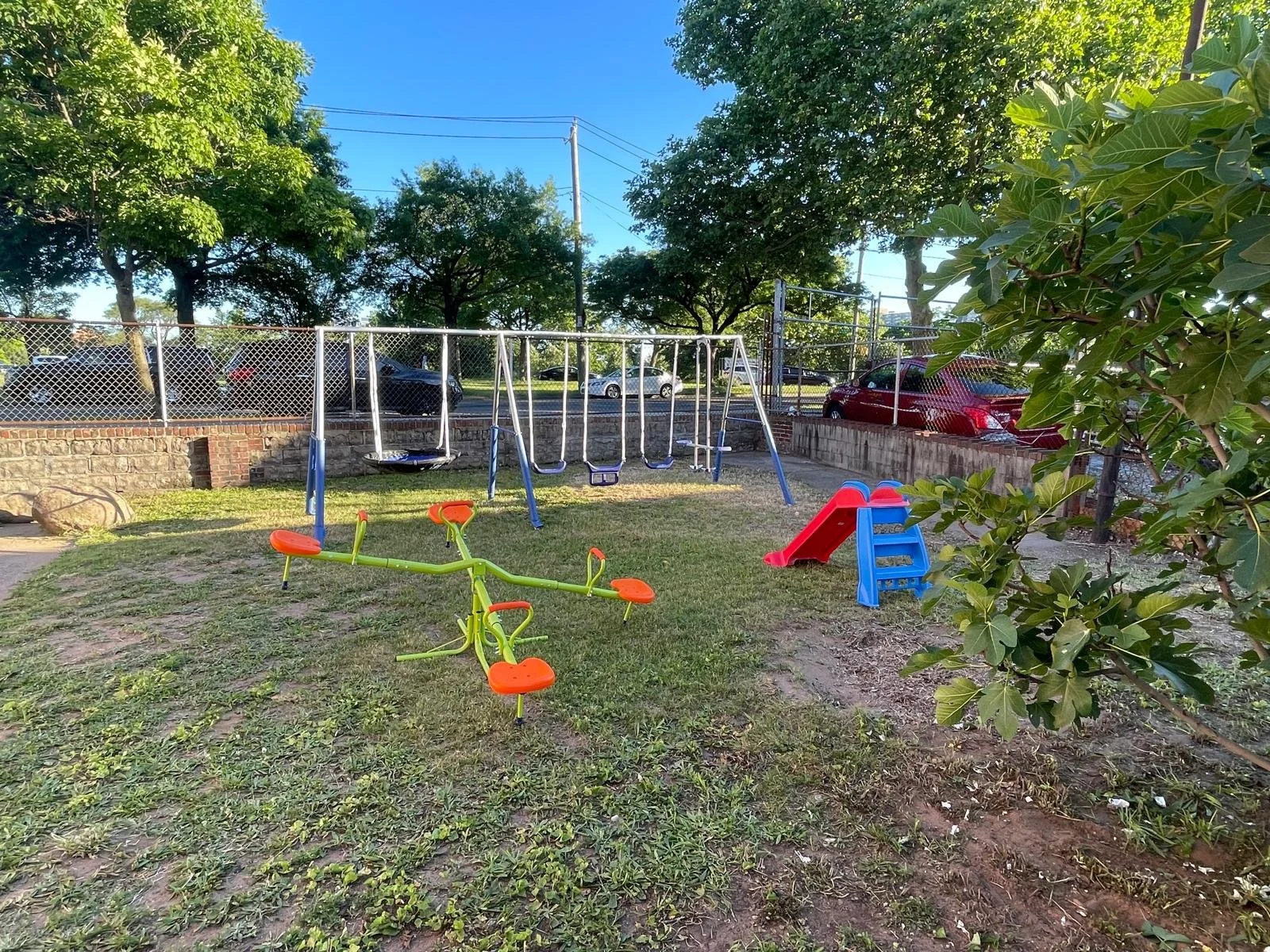 Children's playground with a green and orange see-saw, a red slide, and swings in a grassy area, surrounded by trees and a chain-link fence.
