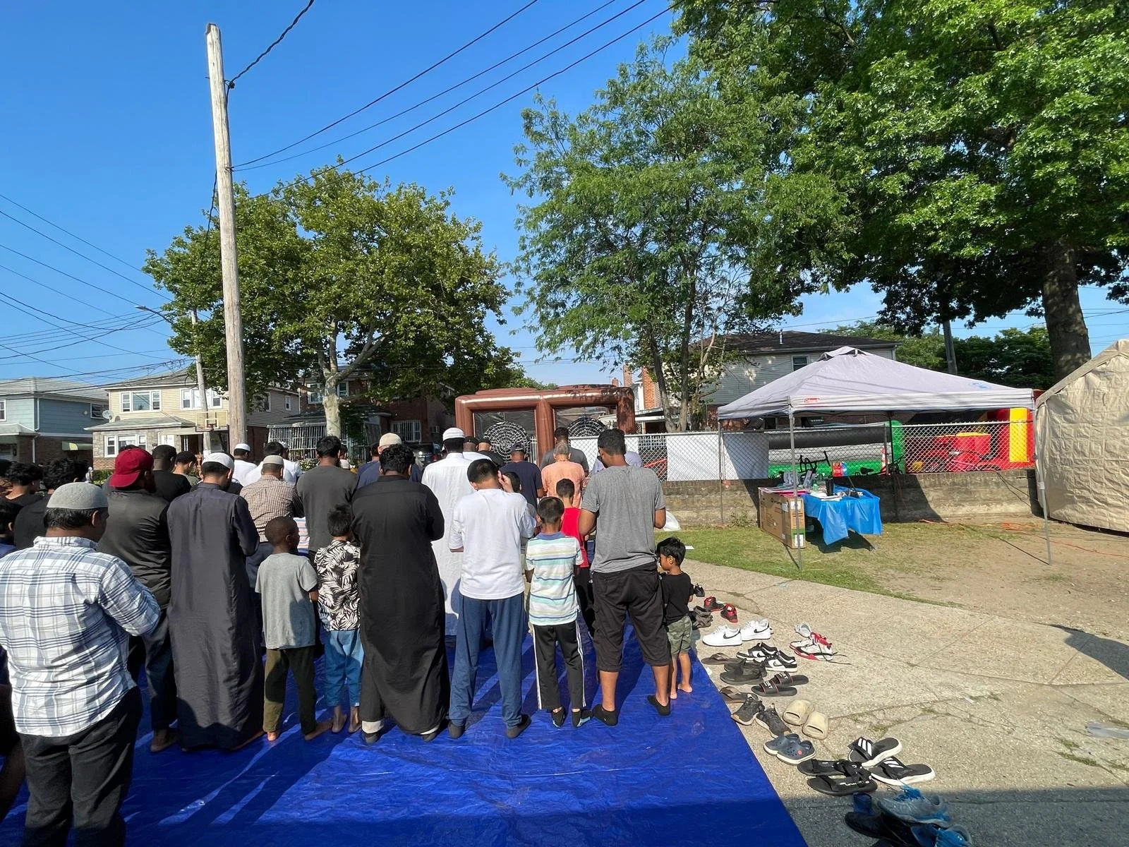 A crowd of people gathered outdoors on a blue tarp, participating in a religious prayer or gathering. Shoes are lined up on the side. Trees, houses, and tents are visible in the background on a sunny day.