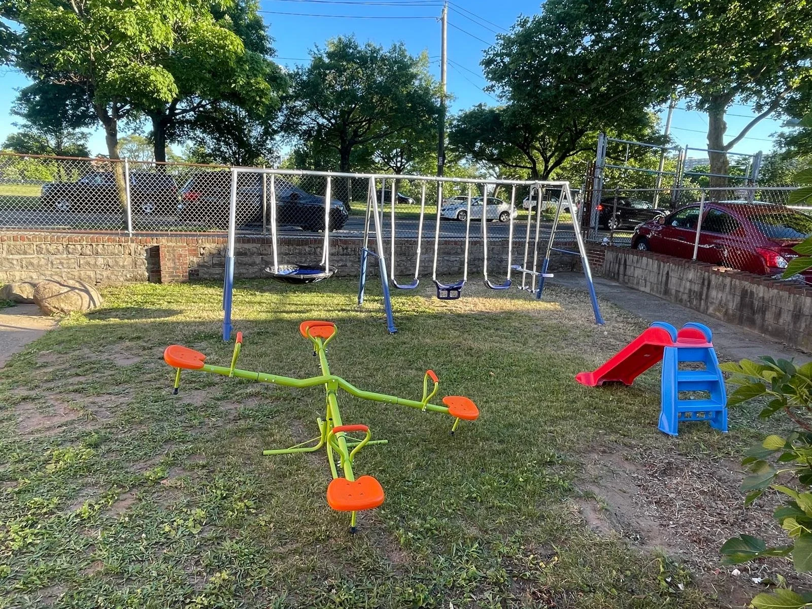 Children's playground with swings, a slide, and a see-saw on grassy area surrounded by trees and parked cars in background.