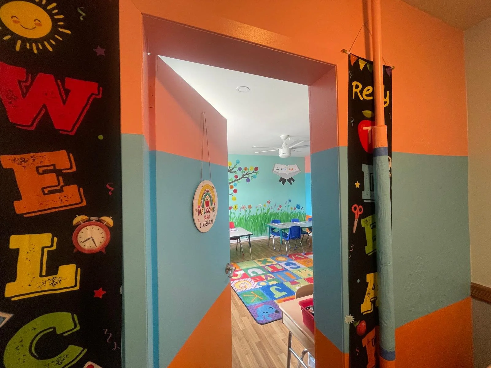 View into a colorful classroom with a bright mural of a tree and flowers on the wall, children's tables and chairs, a rainbow-themed rug with alphabet letters, and playful decor including a cloud and rainbow welcome sign.