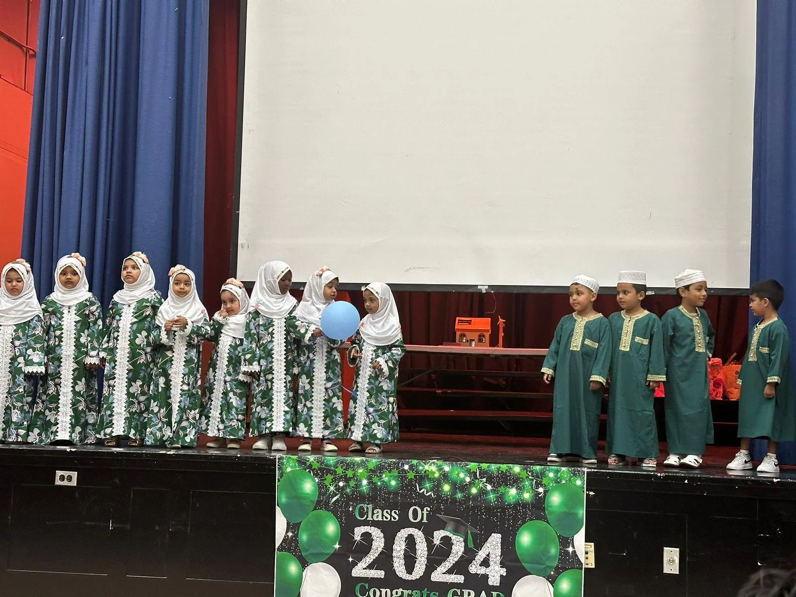 Children in green and white traditional attire on stage with a "Class of 2024" banner.