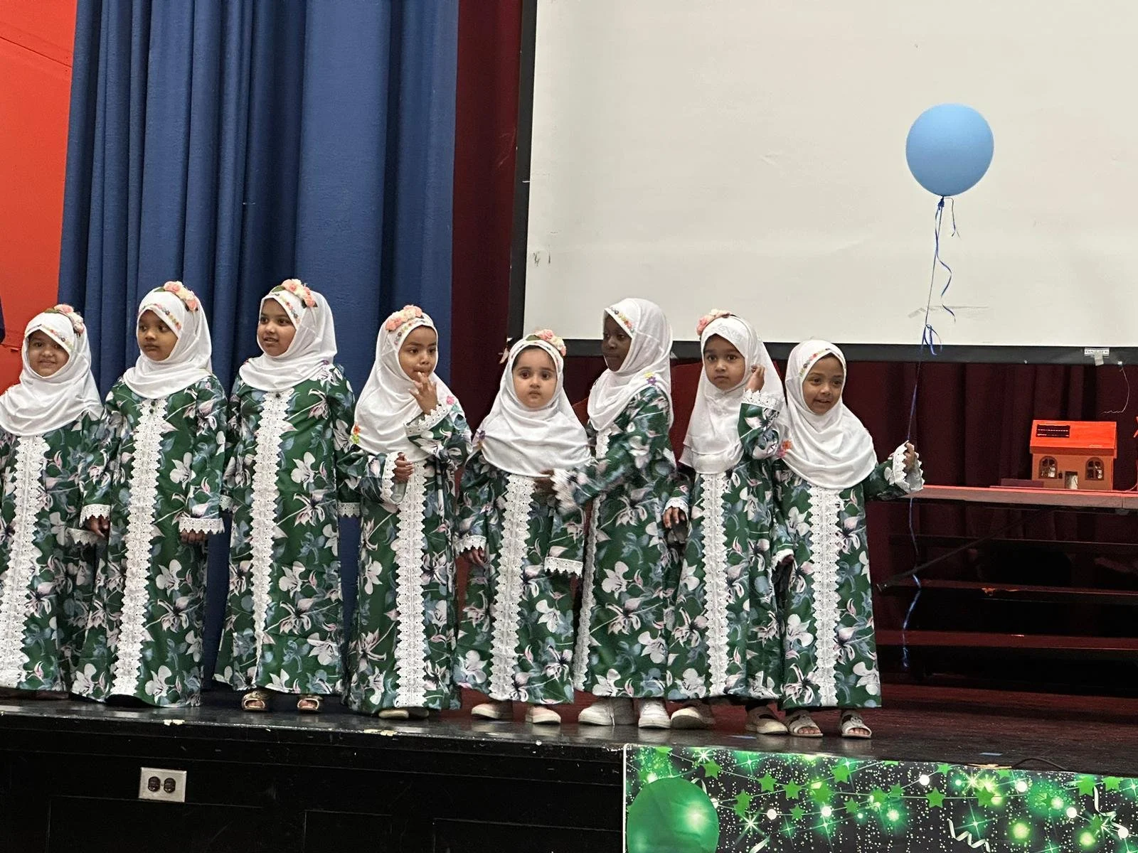 A group of young girls standing on stage wearing matching green floral dresses with white lace and headscarves, holding a blue balloon.