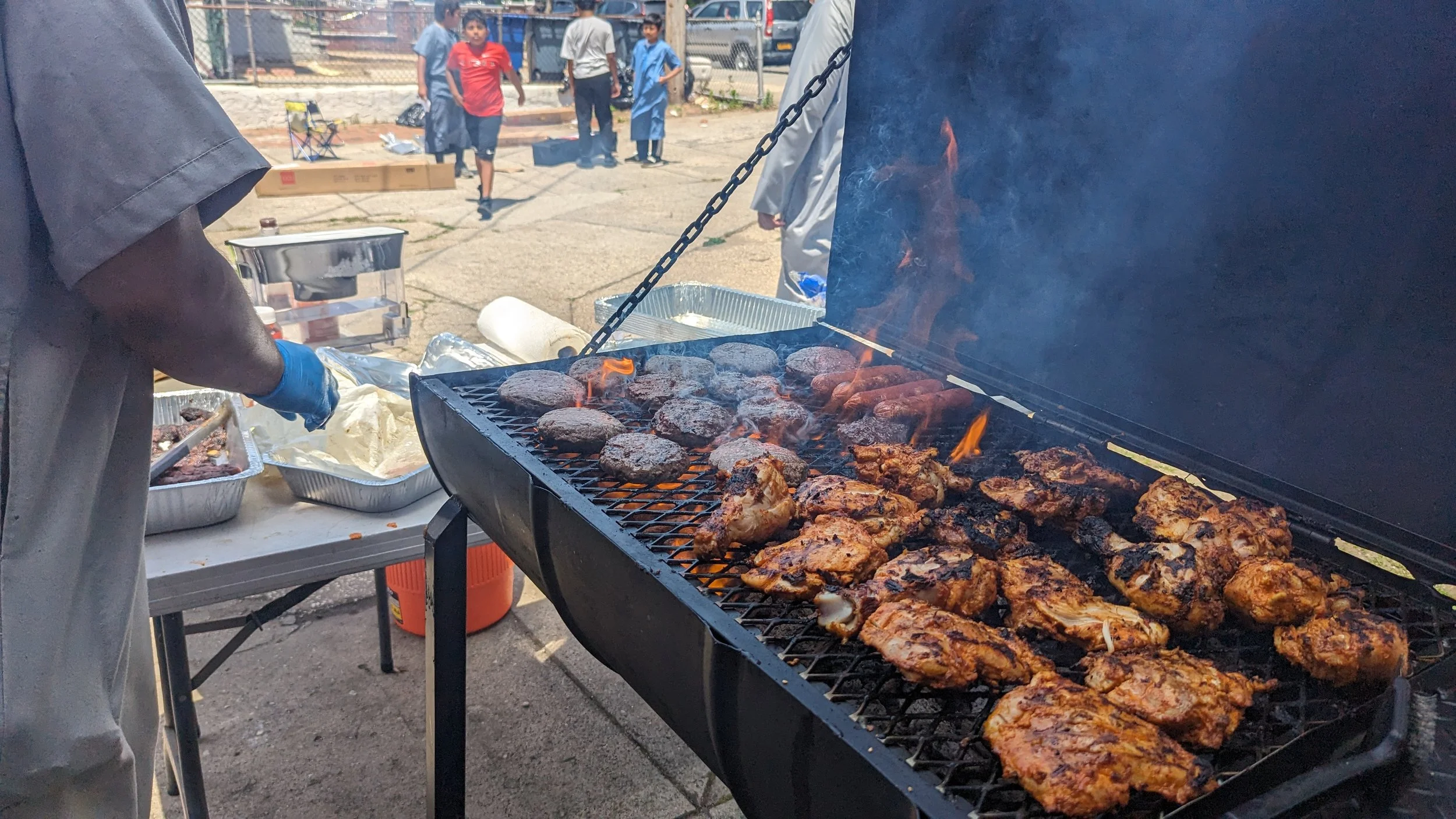 Barbecue grill with seasoned chicken and hamburger patties cooking outdoors, with people in the background.