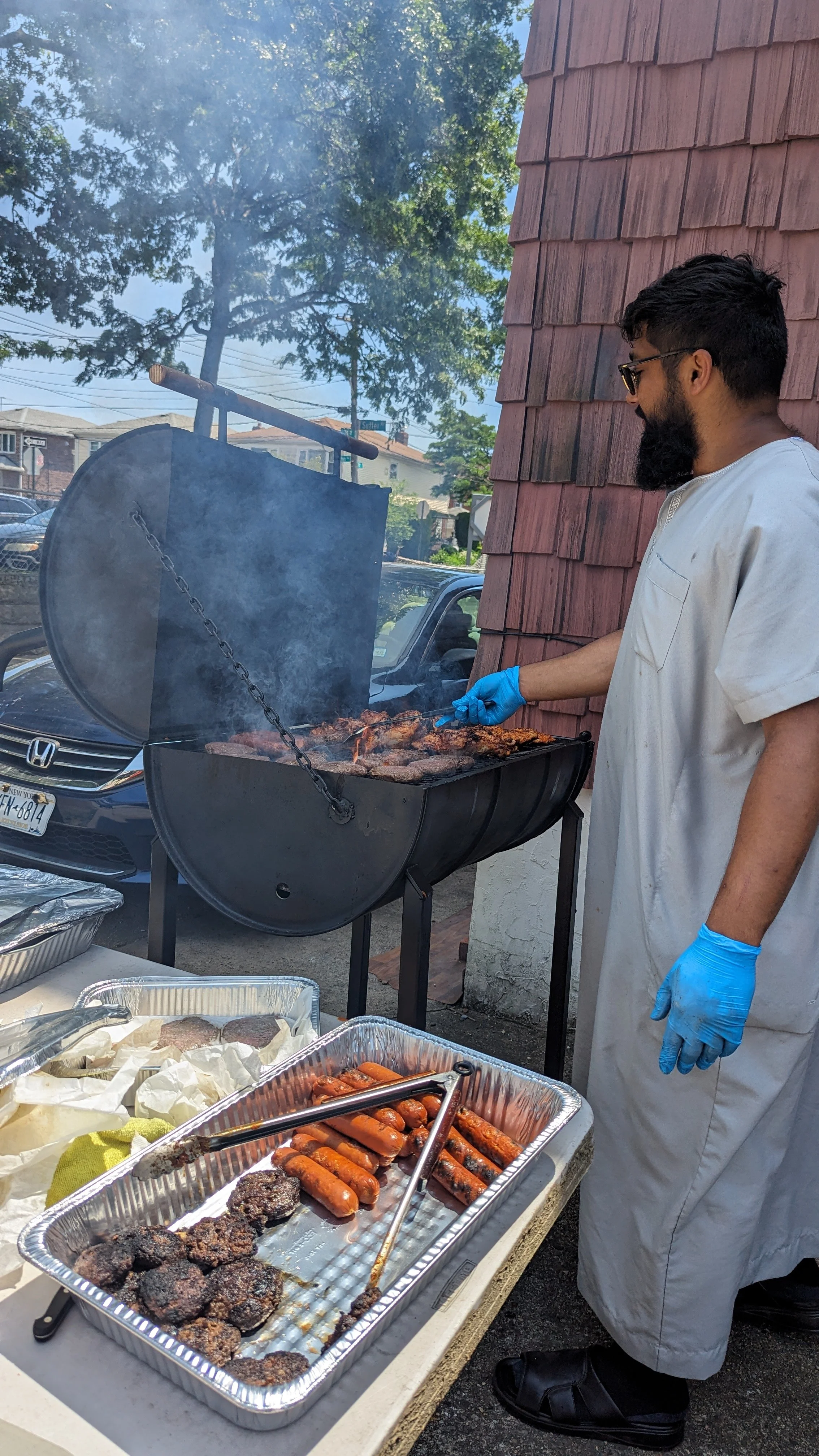 Man wearing a white apron and blue gloves grilling sausages, chicken wings, and skewers on a barbecue grill outdoors, with aluminum trays of cooked food on a table nearby.